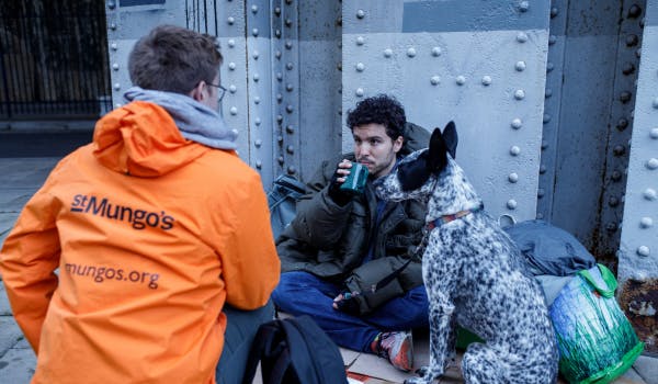 A person in an orange jacket with “St Mungo’s” on the back talking to someone sitting on the ground with a dog nearby.