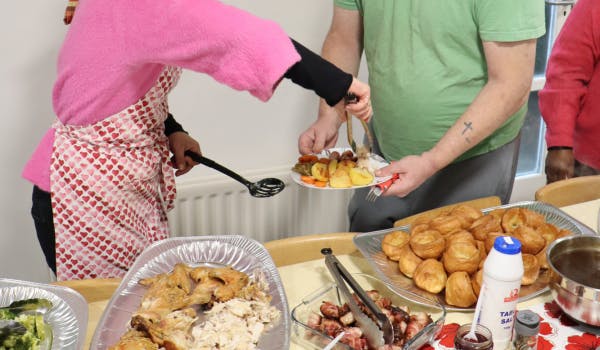 Serving food onto a plate from trays filled with roast meat, vegetables, and Yorkshire puddings.