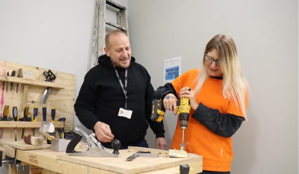 Two people working at a wooden workbench with tools, using a power drill on a piece of wood.