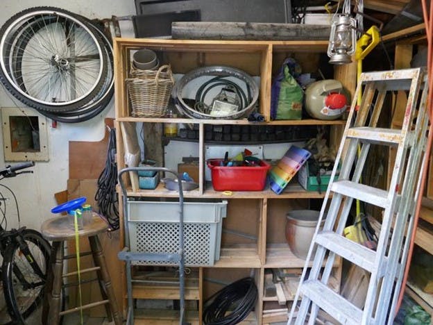 Wooden shelf in a garage is piled with various clutter and surrounded by more mess