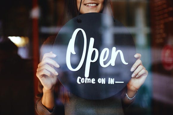 A woman stands in the front window of a business holding a sign that reads “Open. Come on in.”