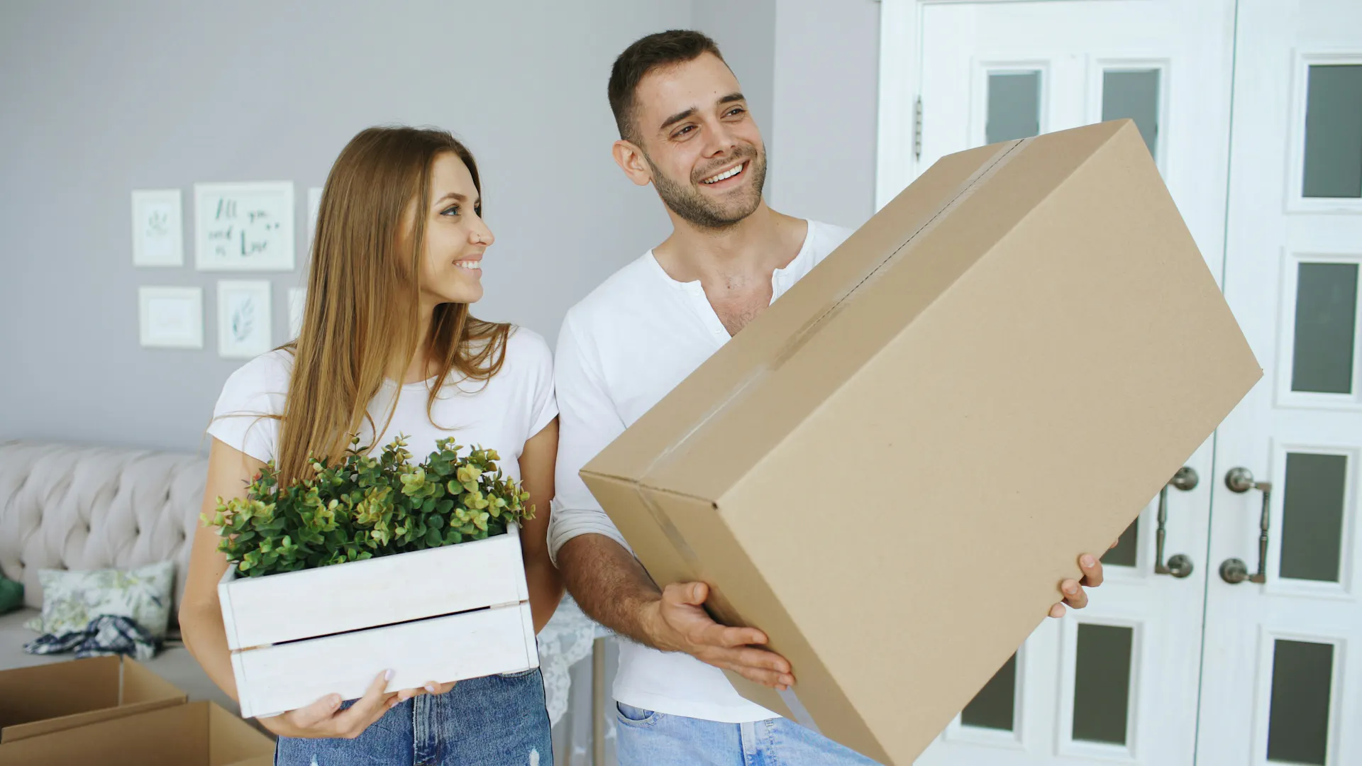 A man and woman carrying a moving box and a small planter box inside of an apartment.