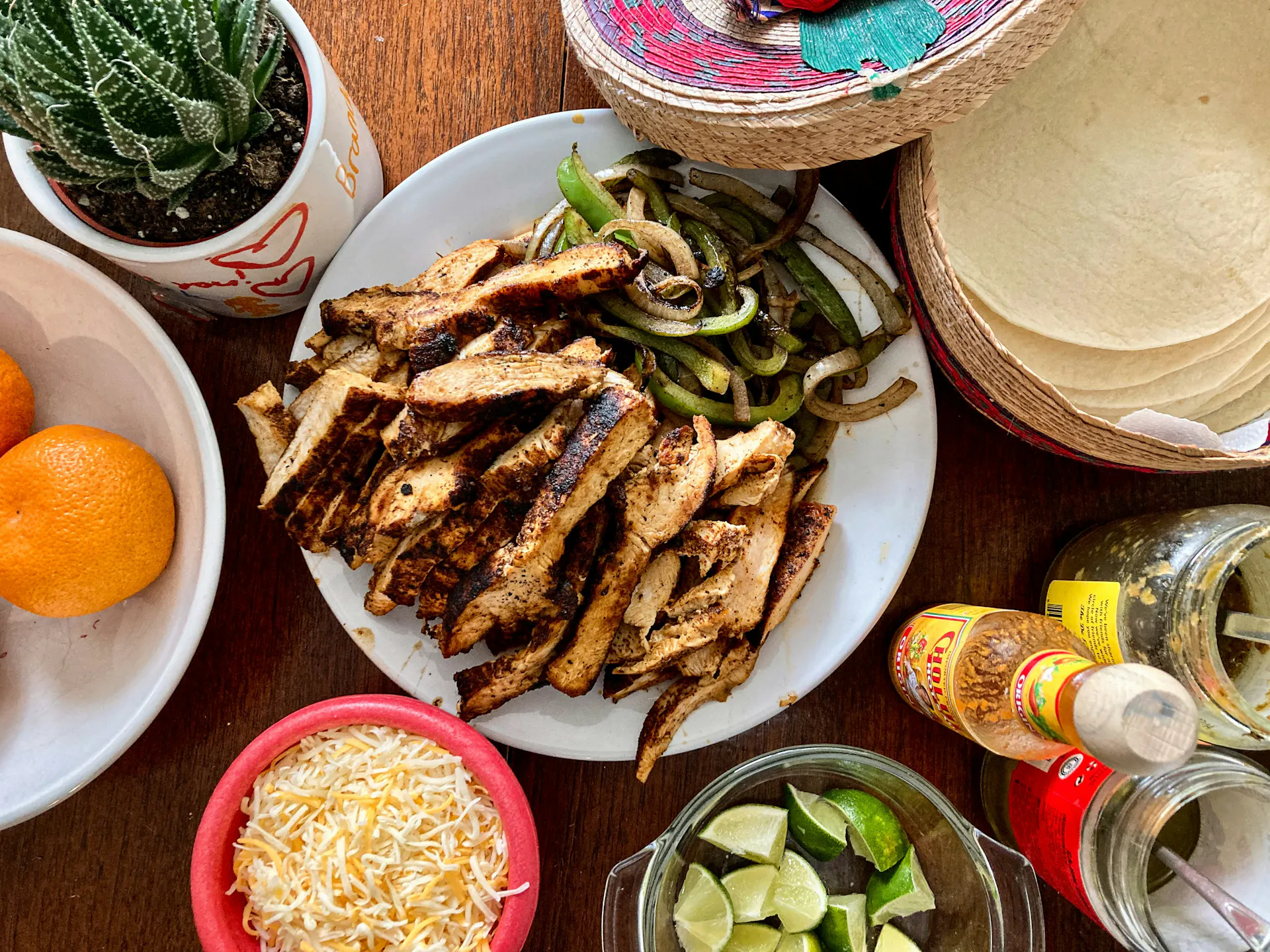A plate of chicken fajitas surrounded by various toppings: cheese, limes, and tortillas.