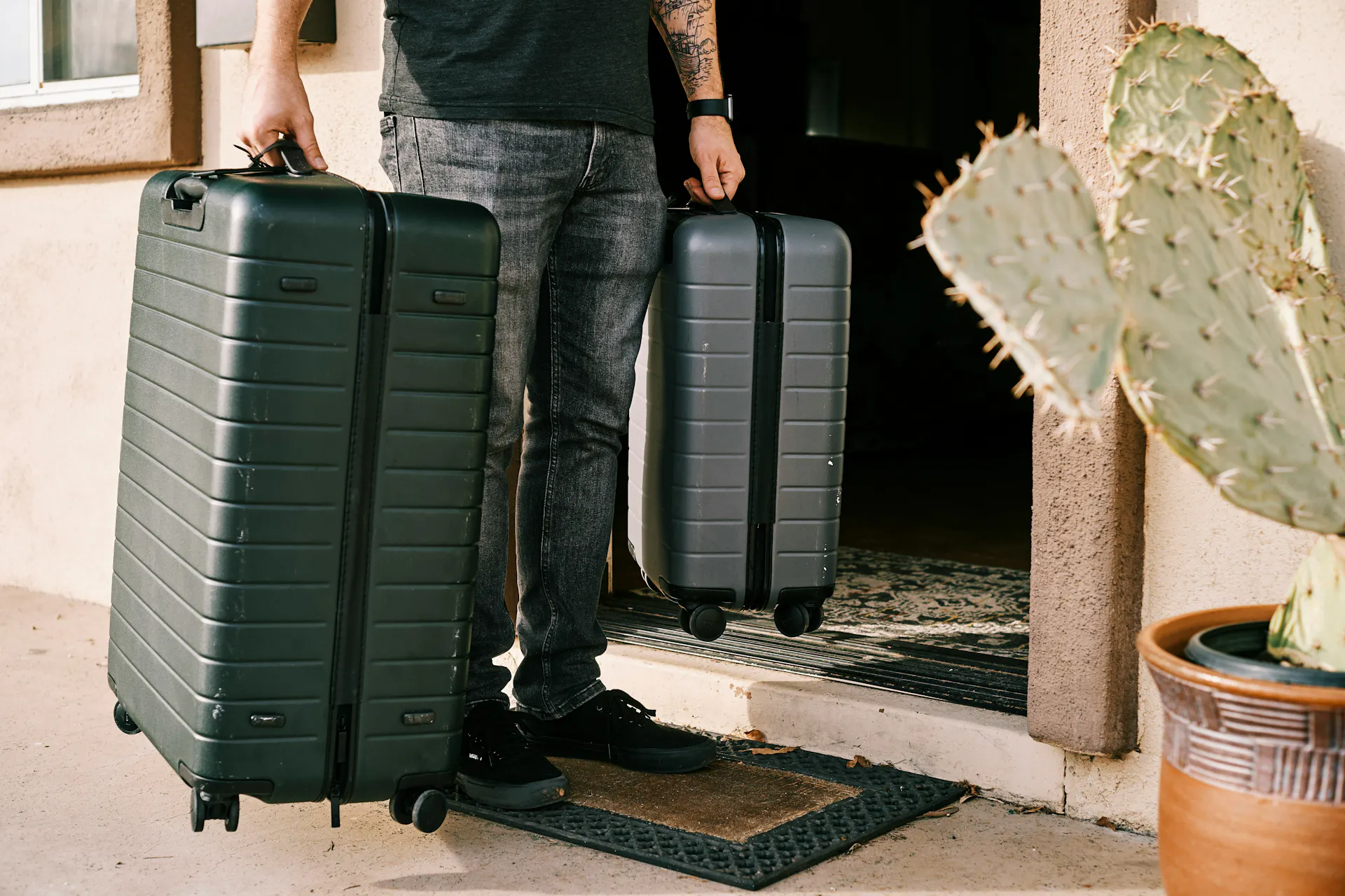 A man in a black shirt and jeans holding two suitcases in the doorway of his new temporary housing.