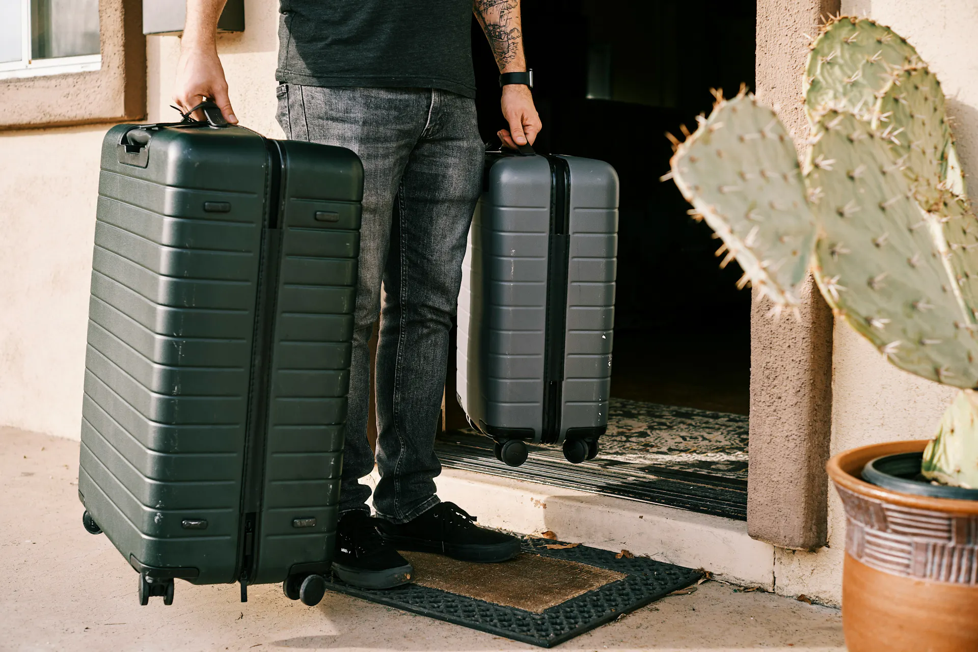 A man in a black shirt and jeans holding two suitcases in the doorway of his new temporary housing.