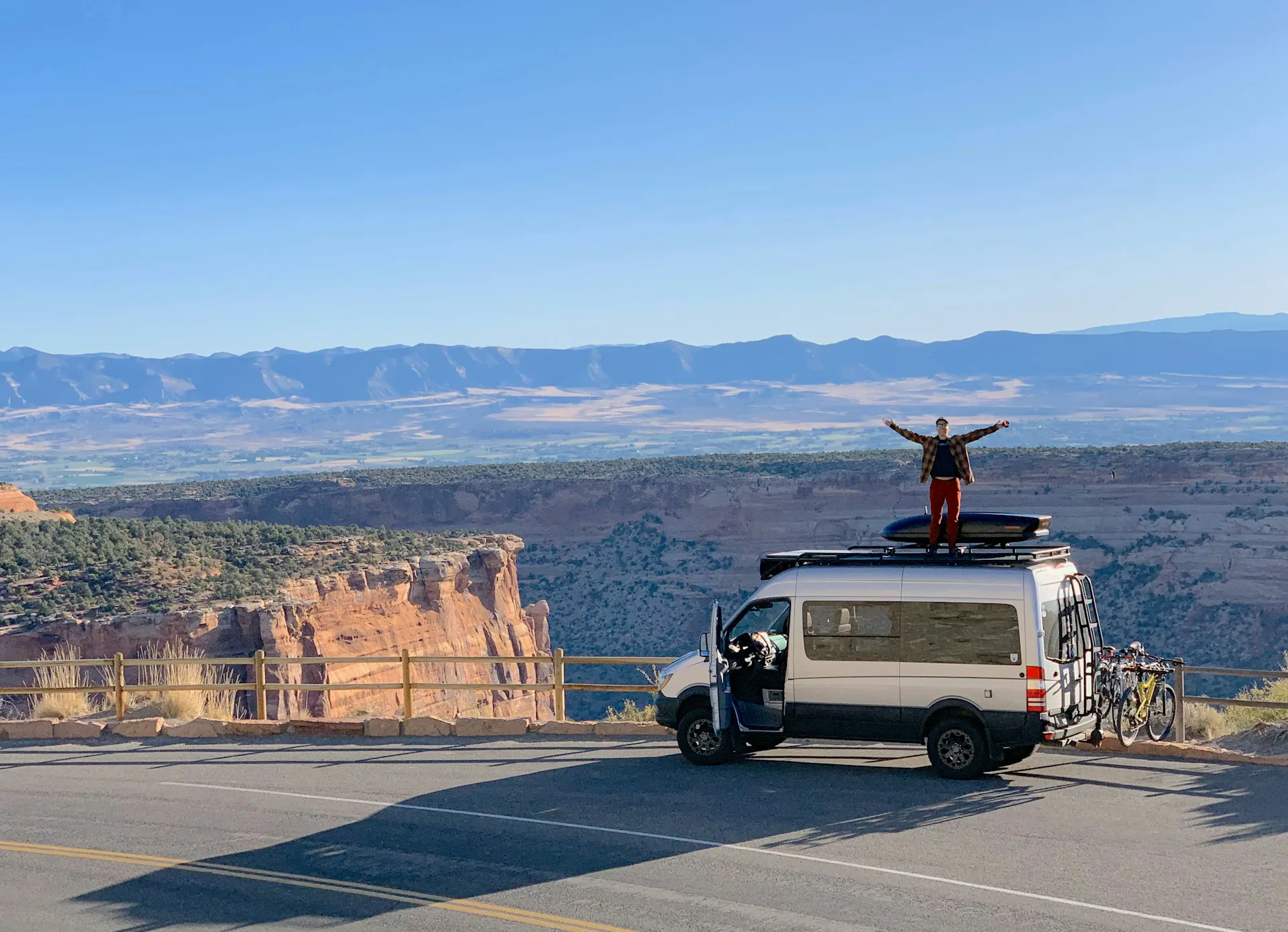 A man standing on his van parked on the shoulder of the highway overlooking mountains out west.