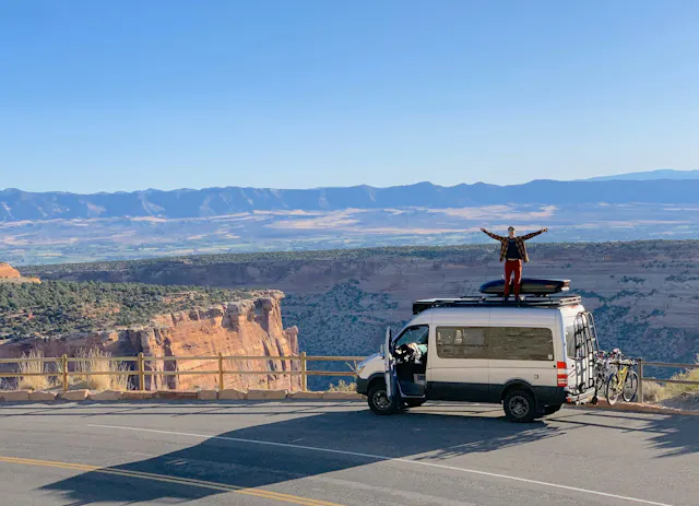 A man standing on his van parked on the shoulder of the highway overlooking mountains out west.
