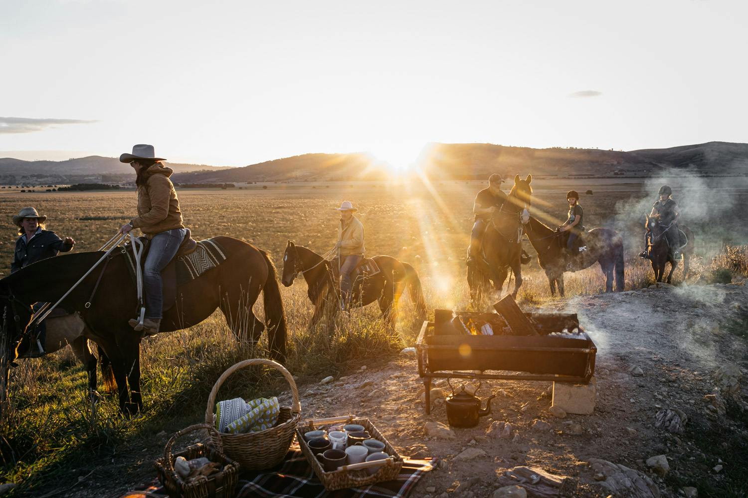 Riders on horseback in the Australian outback with a fire lit and picnic accessories in the foreground and the sun setting over the hills on the horizon