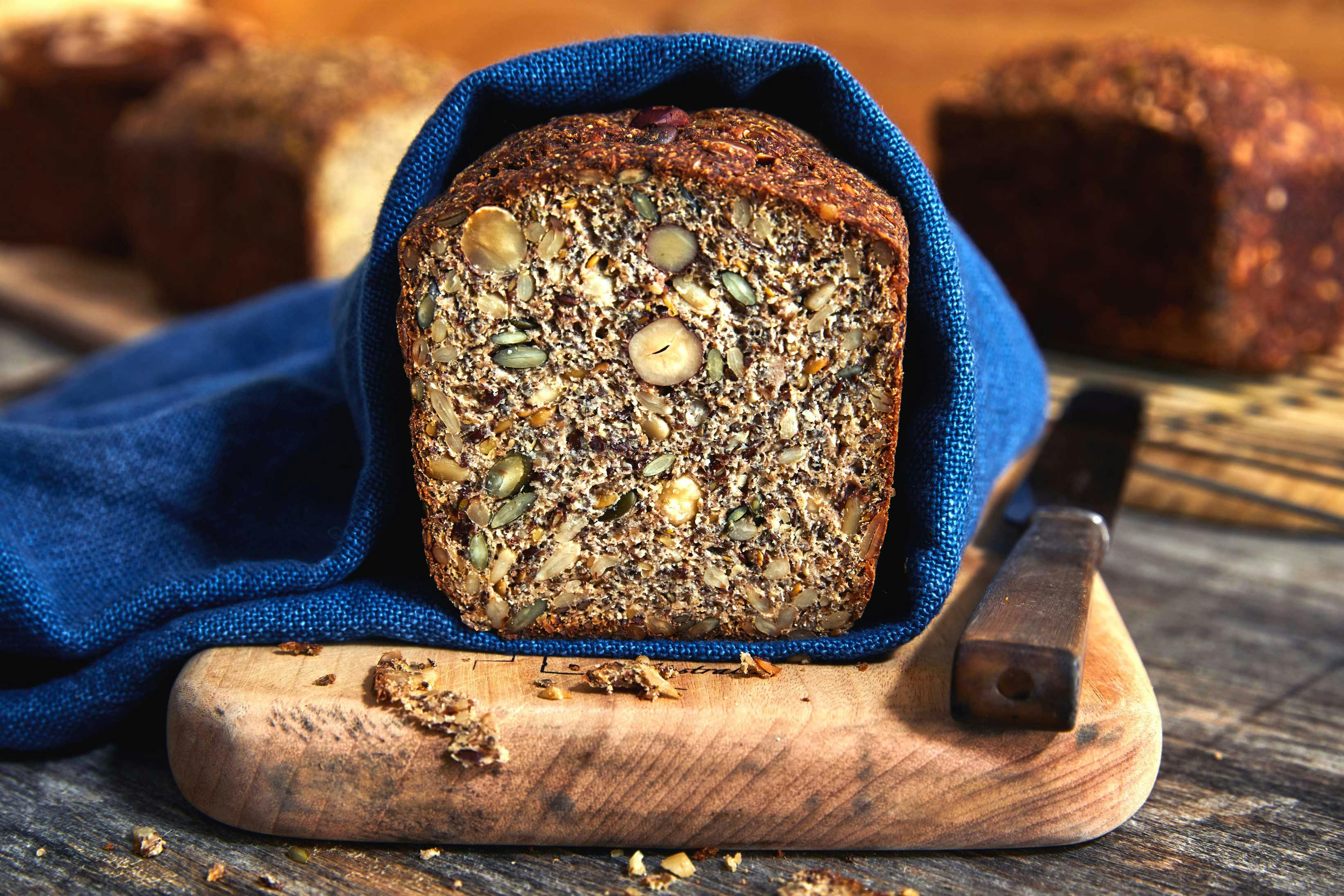 A loaf of cut bread revealing a nutty interior wrapped in a blue napkin on a wooden cutting board with a bread knife adjacent to it