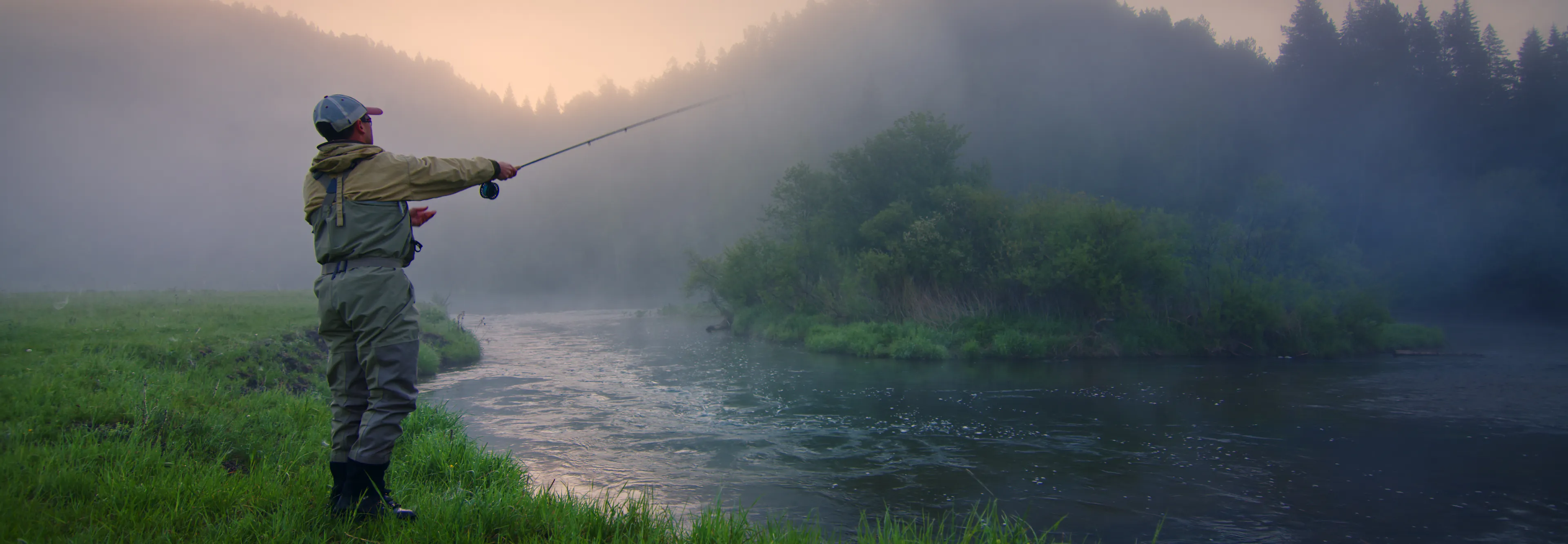 man fly fishing at a river's edge at sunrise with fog rolling