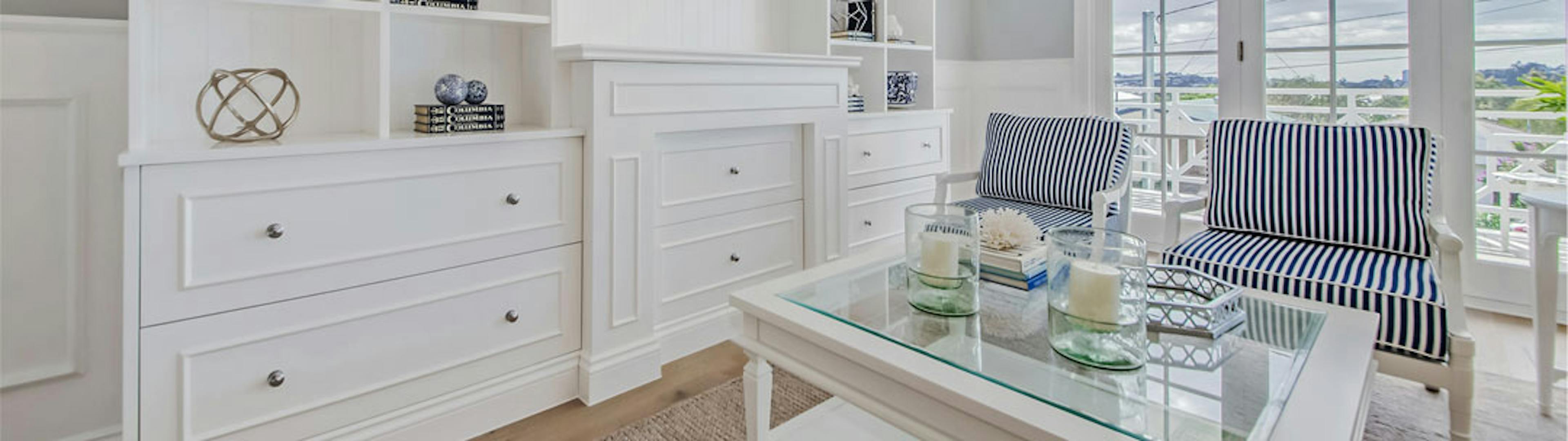 White Hampton style book case that goes from floor to ceiling with storage drawers below open 2pac shelving. Two upholstery chairs near a glass coffee table with blue and white stripe cushions 
