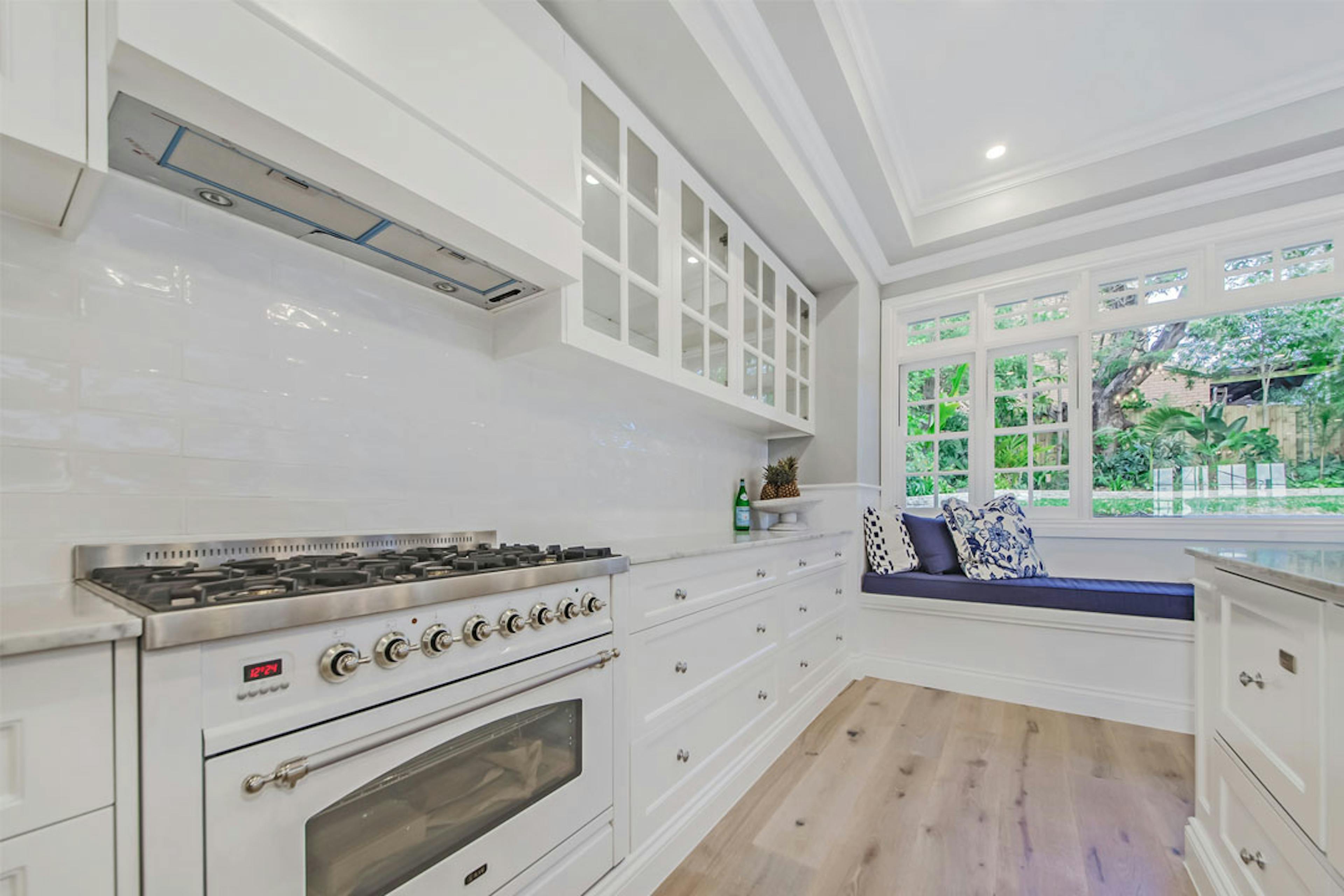 white Hampton style kitchen showing a large oven and range hood. In the background there is views on a garden and a daybed with a dark blue cushion. 