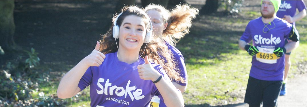 A woman doing a thumbs up, wearing white headphones and wearing a purple t-shirt. She's running and there are a few other women in the background also running.