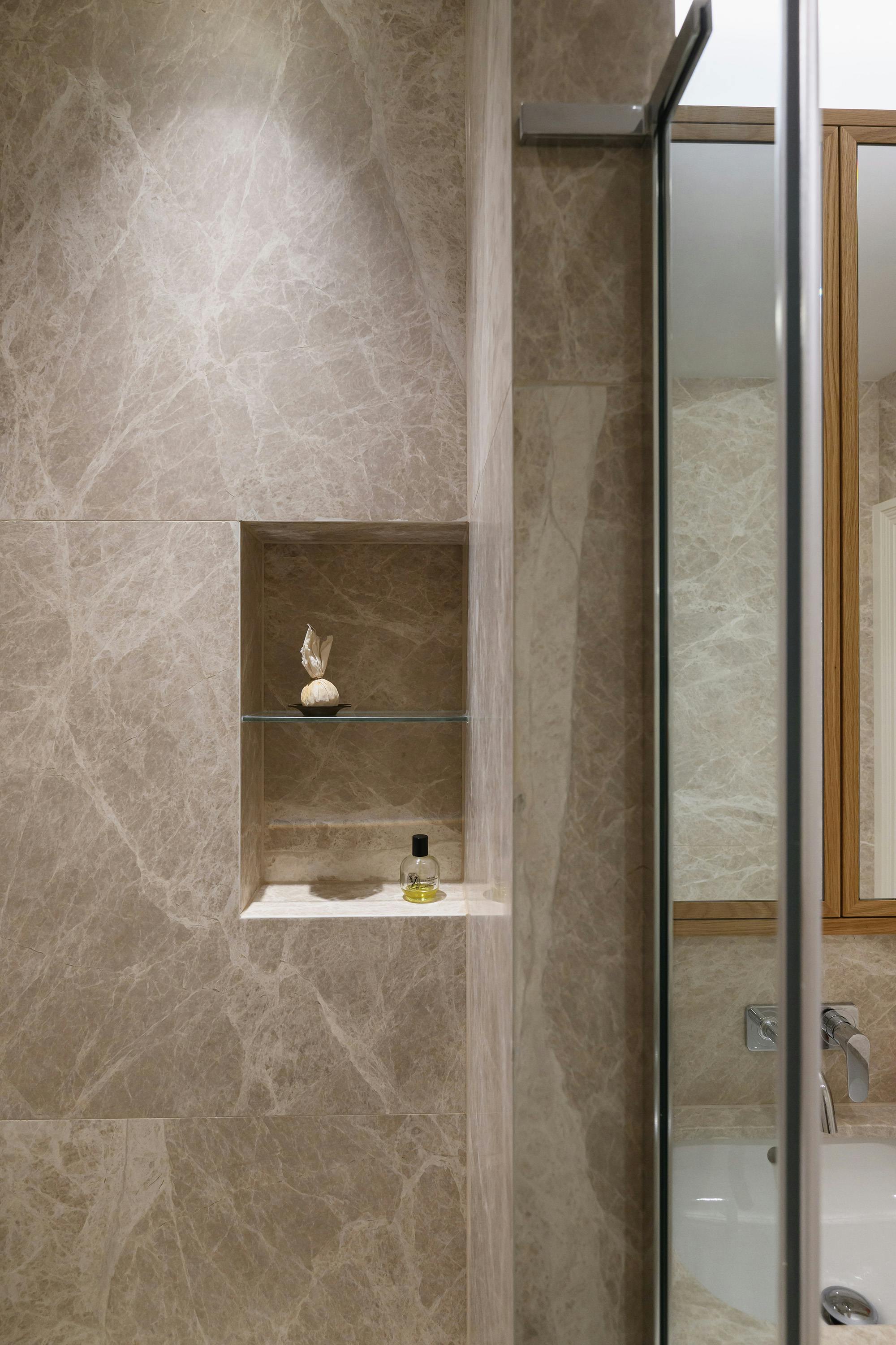 Luxurious bathroom with a walk-in shower enclosed by a glass partition. The walls and built-in shower bench are clad in elegant beige marble with natural veining. A sleek chrome handheld showerhead is mounted on the wall, with a modern concealed mixer tap. A small green glass bottle sits on the shower bench. To the right, a floating wooden vanity with a marble countertop and a matching framed mirror add warmth to the space. The shower floor is tiled in light neutral tones, seamlessly blending with the overall design