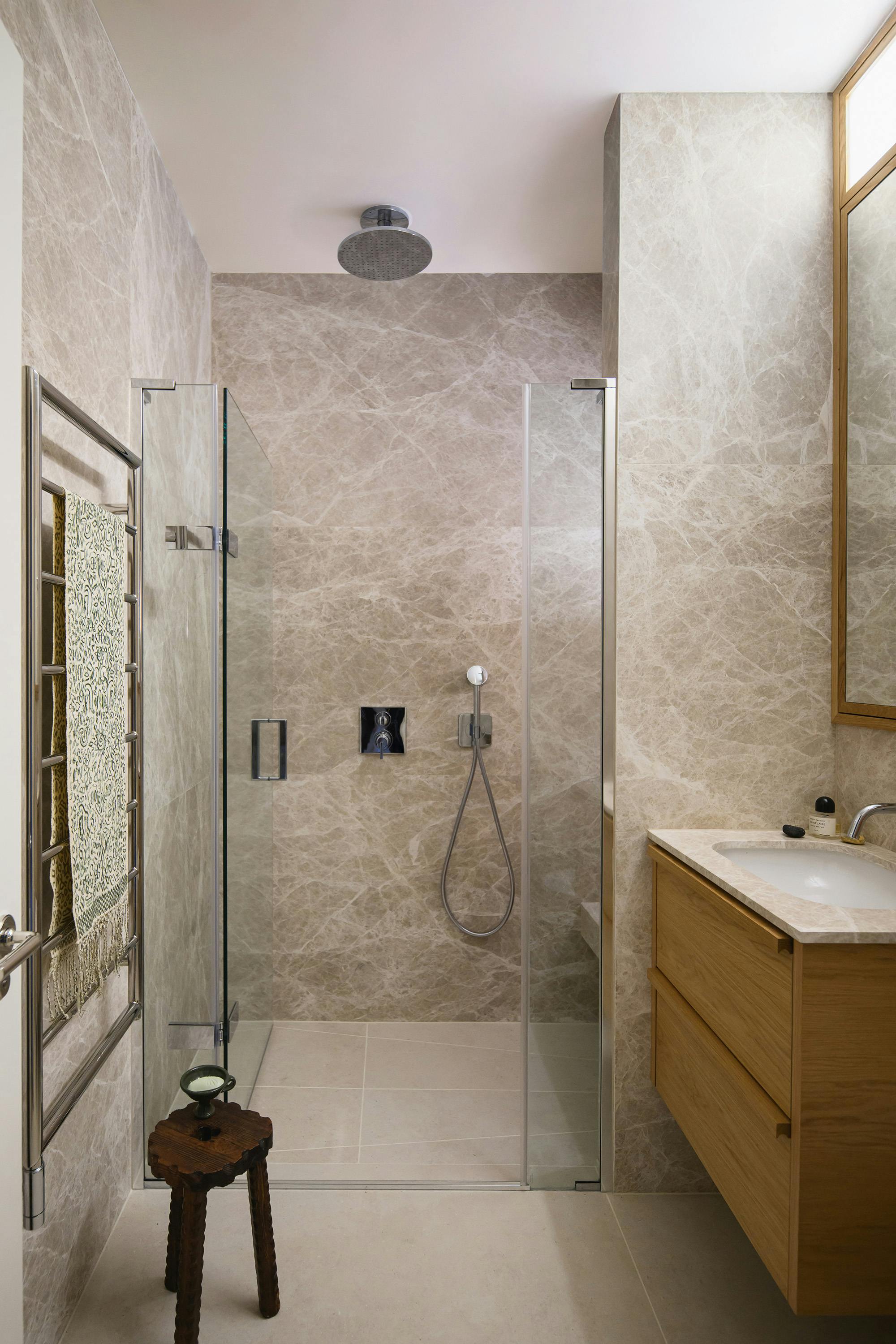 Modern bathroom with a walk-in shower enclosed by glass panels. The walls and floor are clad in beige marble with natural veining. A rainfall showerhead is mounted on the ceiling, accompanied by a handheld shower fixture. To the right, a wooden vanity unit with a marble countertop and integrated sink adds warmth to the space. A wall-mounted mirror with a wooden frame hangs above the sink. On the left, a heated towel rail with a patterned towel is mounted on the wall, and a small rustic wooden stool with a decorative bowl is positioned nearby.