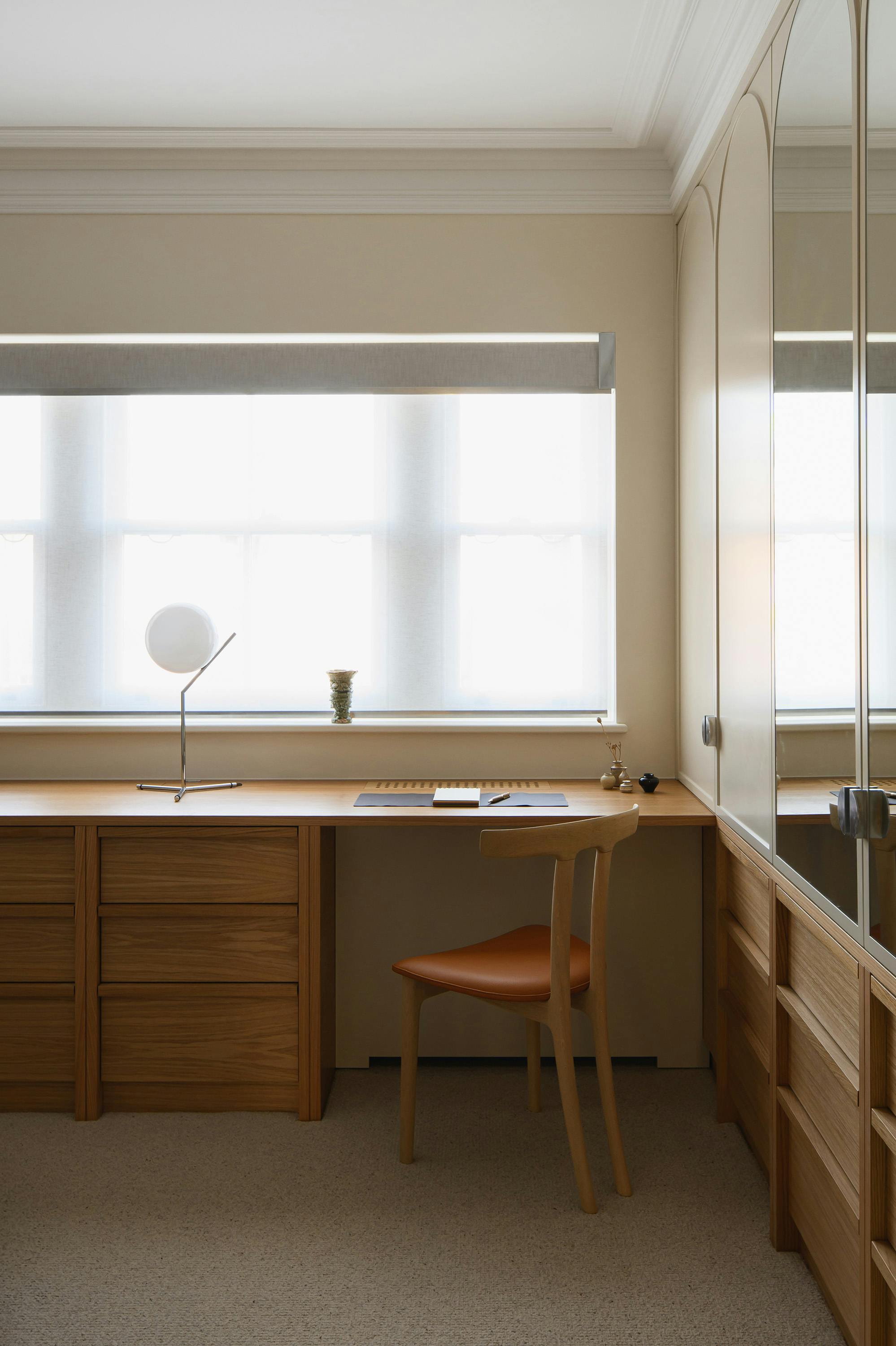 An interior shot of a well-lit room with a classic, elegant design. The room features a light-colored, possibly beige, wall with decorative molding along the top. A large window with sheer white blinds is centered in the frame, providing soft, diffused light. In front of the window is a wooden desk with a minimalist design, featuring a smooth surface and drawers on the left side. A small, white table lamp with a spherical shade is placed on the left side of the desk. A wooden chair with a leather seat is positioned at the desk. On the right side of the room, a large, ornate mirror with arched segments reflects the room, creating a sense of depth and doubling the space. The floor is covered in a light-colored carpet. The room's overall aesthetic is refined and sophisticated, with a focus on natural light and elegant furnishings.