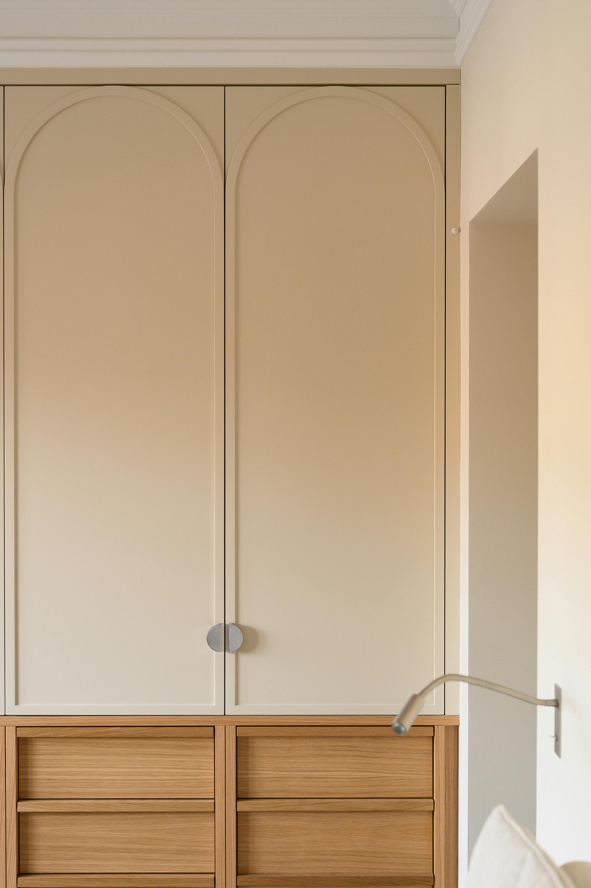 Close-up view of a built-in wardrobe with arched panel doors in a soft beige finish, complemented by round metal handles. Below the wardrobe, there is a set of wooden drawers with a natural oak finish, providing additional storage. A minimalist, wall-mounted reading light extends from the side, positioned near a partially visible white pillow, suggesting a bedside placement. The room has a neutral colour palette, with soft lighting highlighting the elegant detailing of the cabinetry and classic crown moulding along the ceiling.