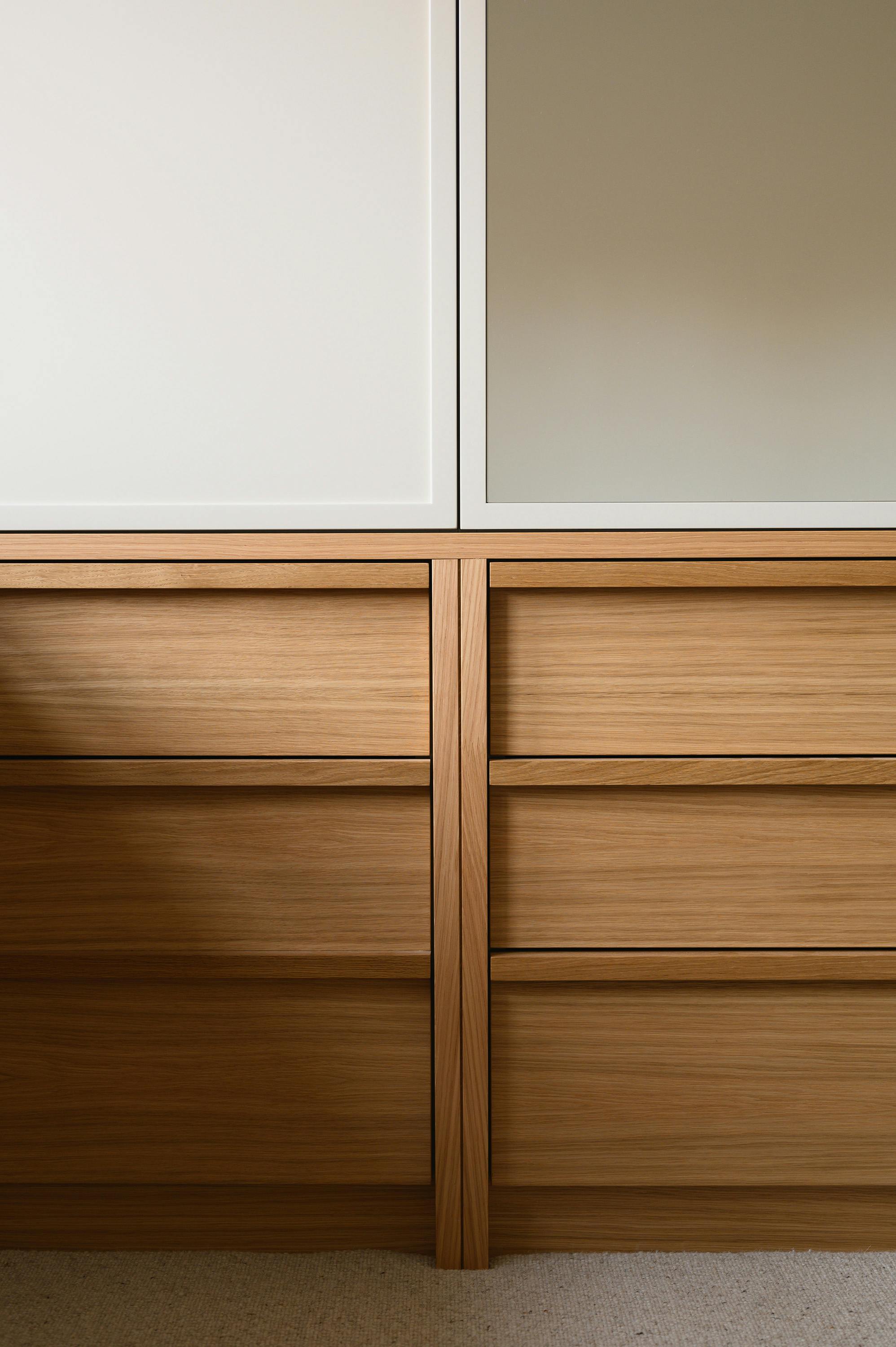 Close-up view of a custom-built wooden storage unit featuring multiple drawers with a natural oak finish. The upper section consists of a white panel on the left and a mirrored panel on the right, framed with clean lines. The combination of wood, white, and mirrored surfaces creates a balanced and modern aesthetic. The lower section showcases the detailed craftsmanship of the wooden drawers, with recessed handles and a symmetrical design. A soft, textured carpet is visible at the bottom of the image, adding warmth to the space.