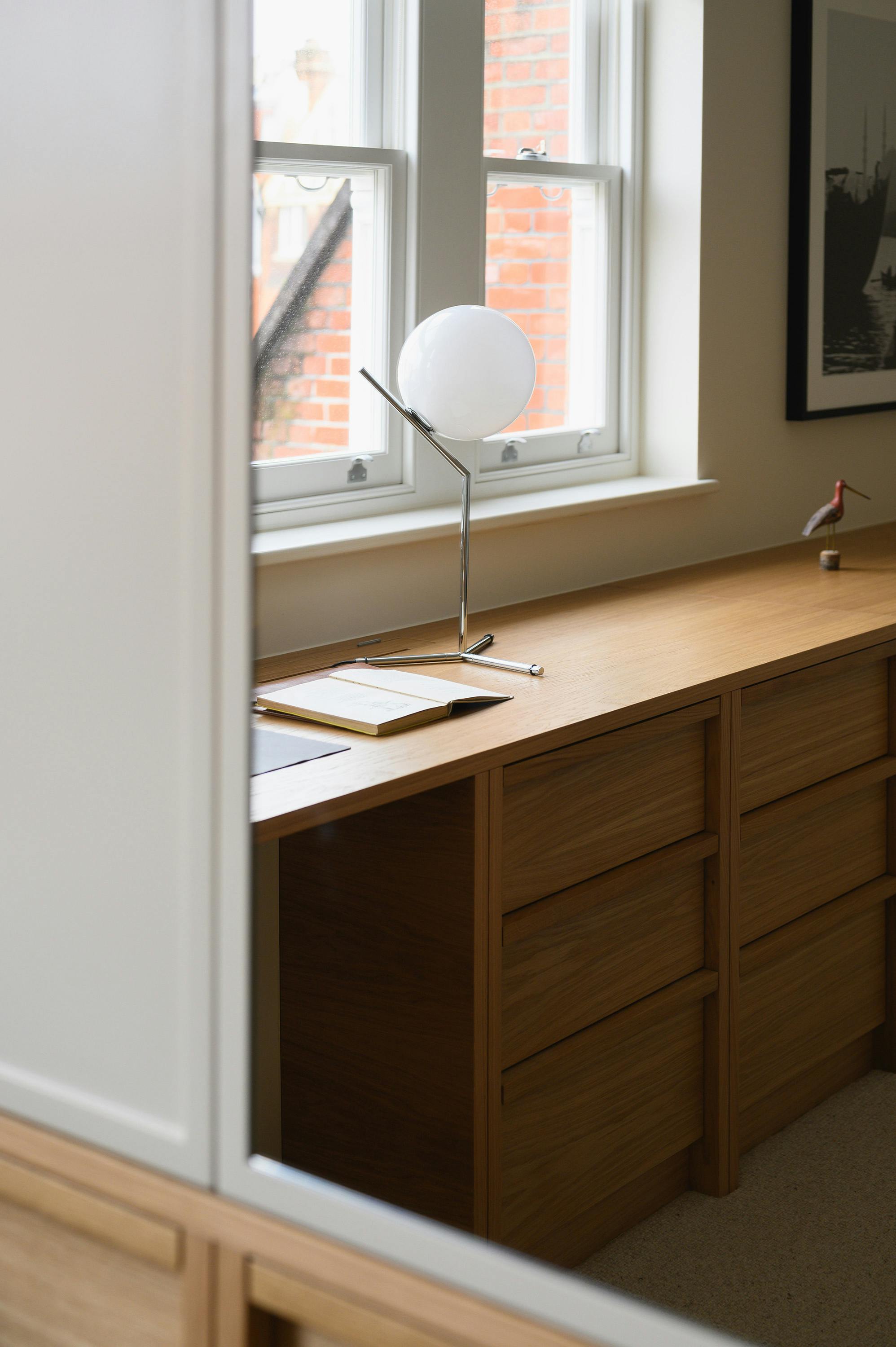 Close-up view of a wooden built-in desk with multiple drawers, positioned beneath a set of white-framed windows that allow natural light to stream in. On the desk, a modern table lamp with a spherical white glass shade and a sleek chrome stand is placed next to an open notebook. A framed black-and-white artwork hangs on the wall in the background, and a small decorative wooden bird figurine is perched on the far end of the desk. The reflection in a nearby mirror reveals part of a wardrobe with wood and cream-colored panelling, adding to the warm, neutral aesthetic of the room.