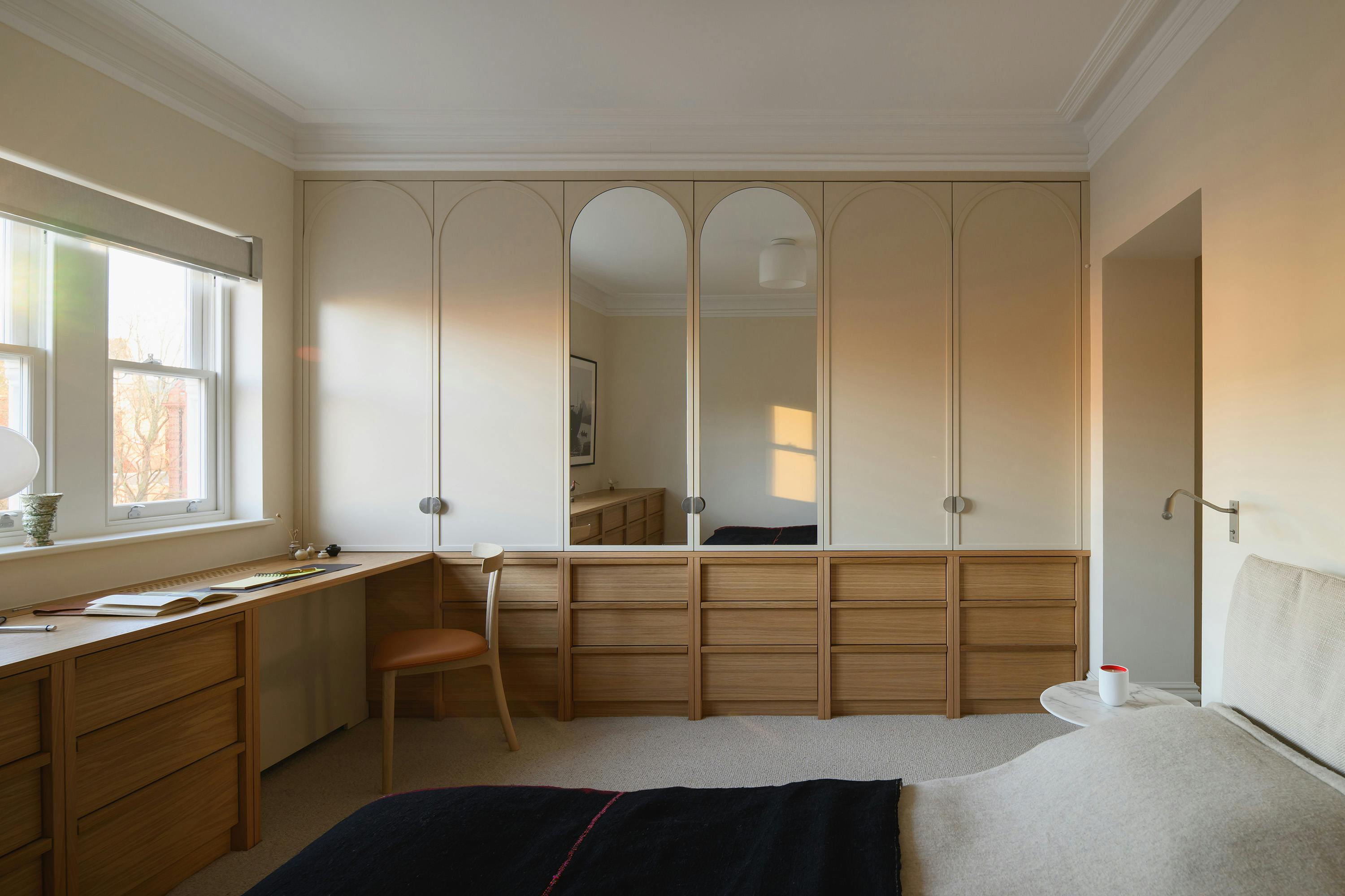 A modern bedroom with a warm, neutral colour palette featuring custom-built wooden storage units and arched wardrobe doors. The built-in desk extends along the window, with an open book, a lamp, and a chair with a leather seat. The wardrobe has elegant arched panelling, with two mirrored doors reflecting the room's interior. A neatly made bed with a soft beige upholstered headboard and a black throw is visible in the foreground. The soft glow of natural light highlights the textures of the wood, fabric, and carpet, creating a serene and inviting atmosphere.