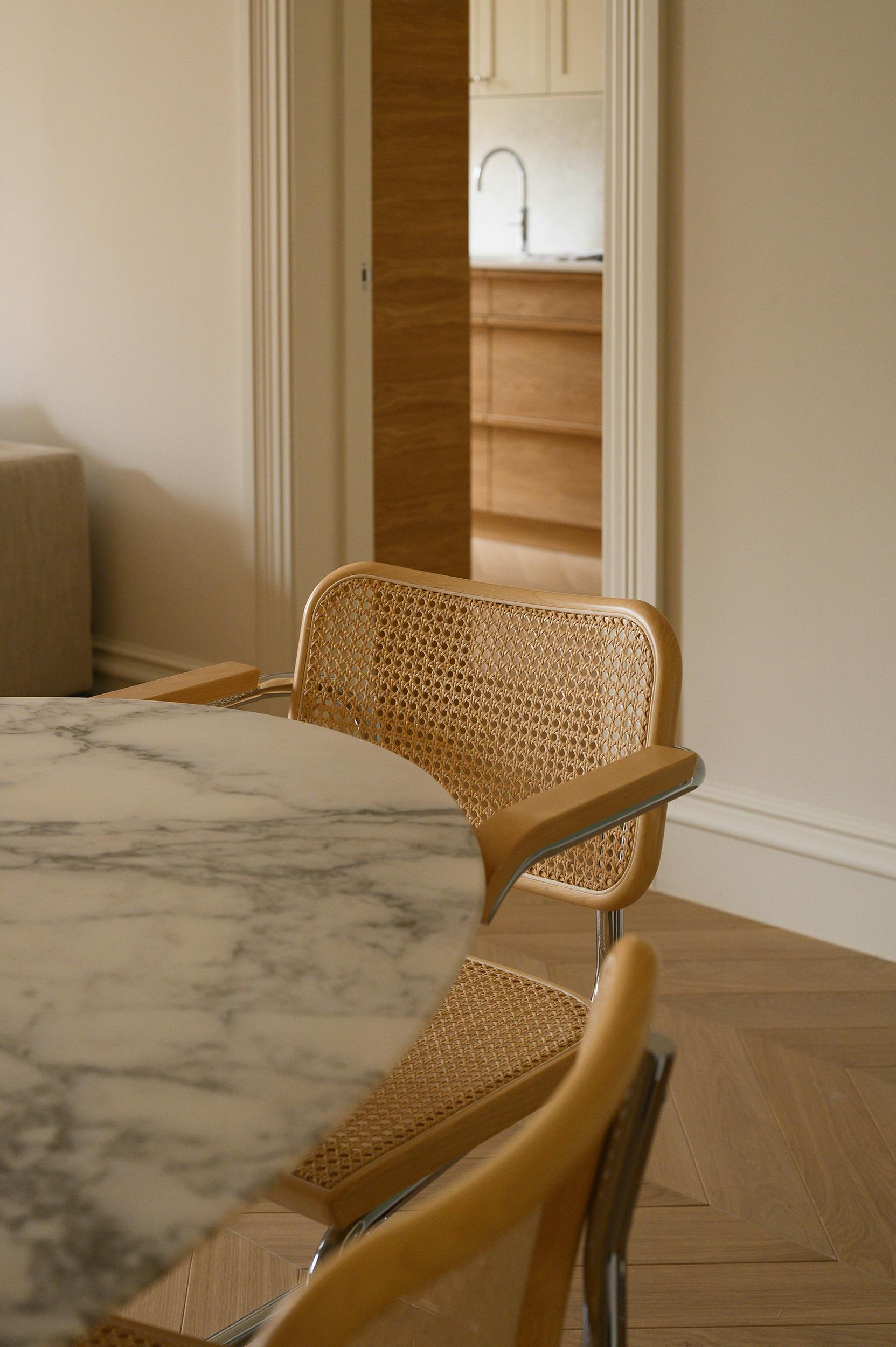 A close-up view of a dining area featuring a round marble table with a veined pattern and woven cane chairs with a light wood frame and chrome detailing. The chairs have a mid-century modern aesthetic, with a curved backrest and armrests. In the background, an open doorway leads to a kitchen with wooden cabinetry, a stainless steel faucet, and a light-coloured backsplash. The room is illuminated by natural light, highlighting the warm tones of the wooden flooring and furniture.