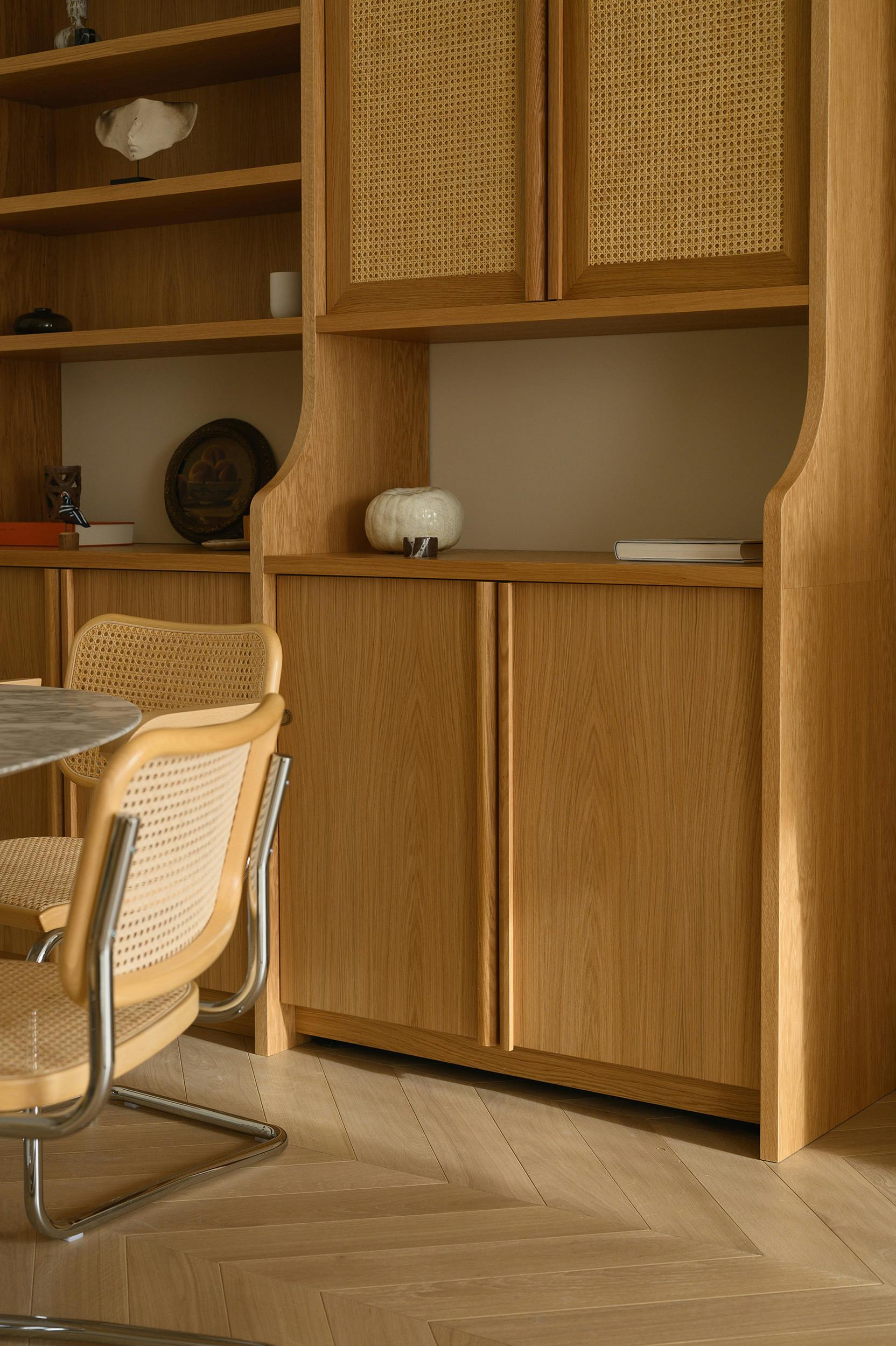 A close-up of a dining area featuring a wooden built-in cabinet with woven cane panel doors and open shelving, adorned with decorative objects such as a ceramic vase, a framed artwork, and small sculptures. In the foreground, a round marble table is partially visible, accompanied by classic mid-century modern cantilevered chairs with woven cane seats and backs, chrome frames, and wooden armrests. The light oak herringbone flooring complements the warm, neutral tones of the furniture and decor.