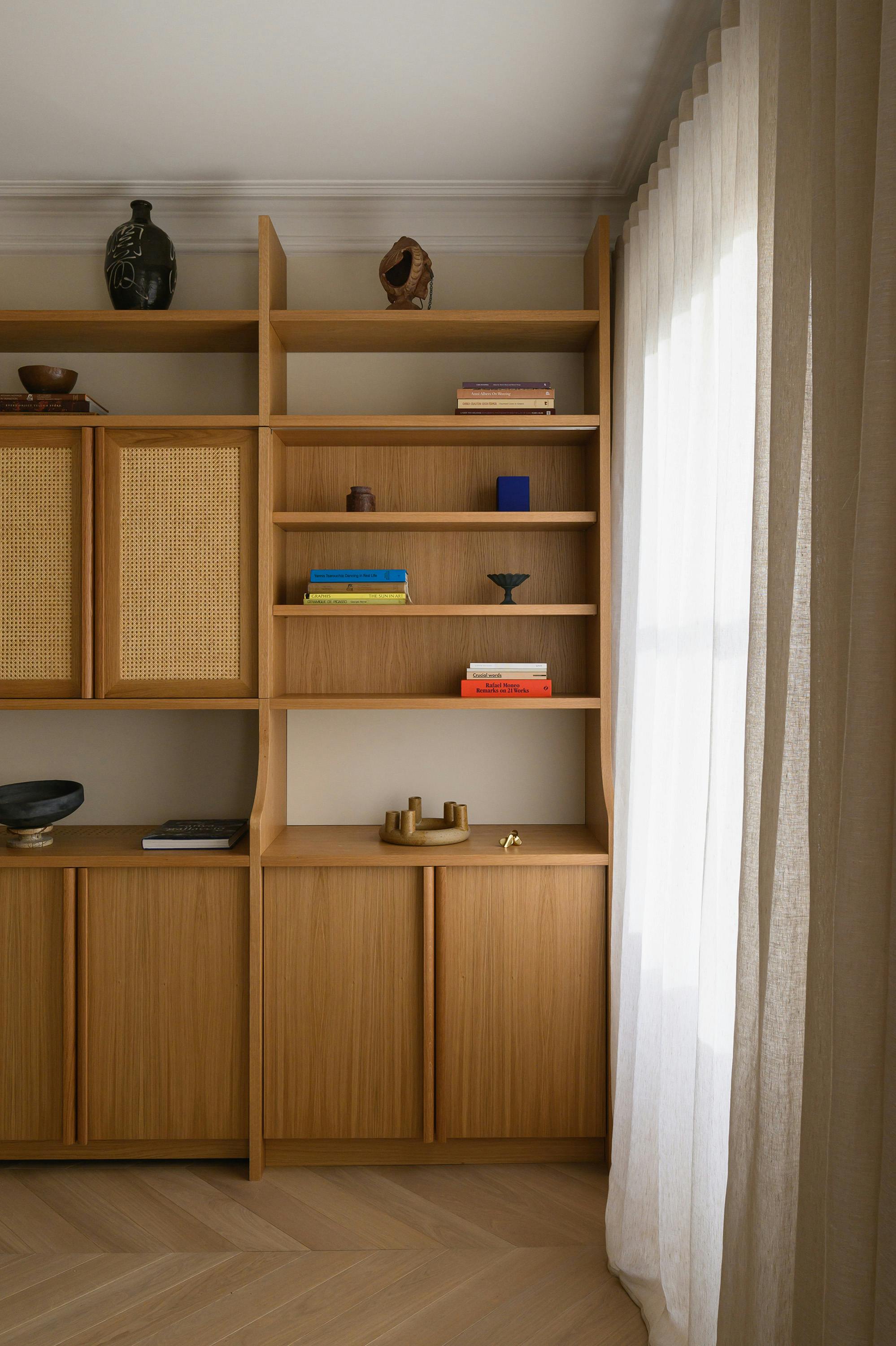 A built-in wooden shelving unit extending to the ceiling, featuring open shelves and cane-panelled cabinets. Various books, ceramics, and decorative objects are arranged on the shelves. A large window with sheer linen curtains allows natural light to filter in, complementing the warm wood tones and chevron-patterned wooden flooring.