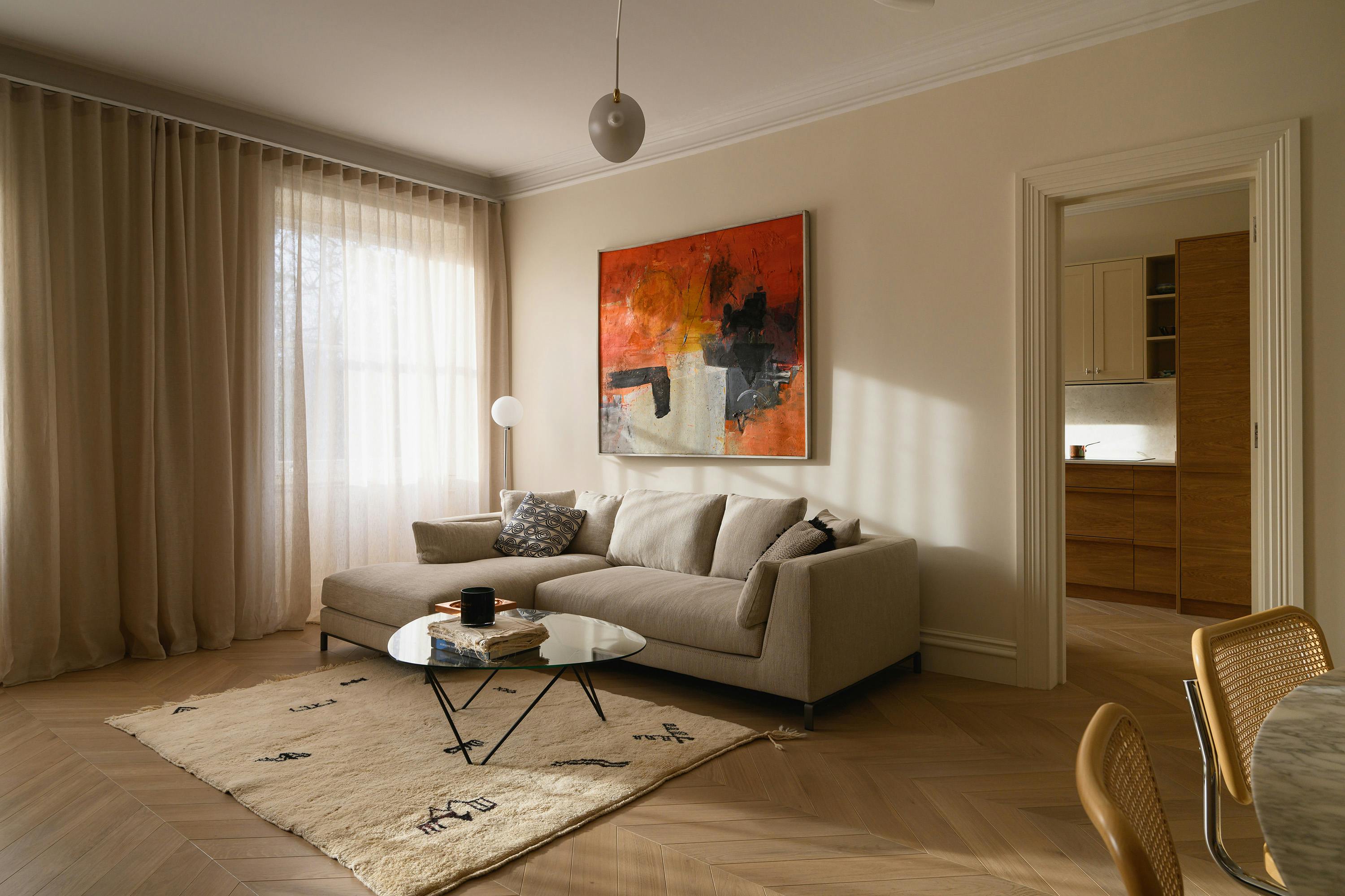 A cozy living room with a neutral-toned sectional sofa adorned with patterned and textured cushions. A modern black metal and glass coffee table sits atop a cream-colored, abstract-patterned rug. A bold, abstract painting in shades of orange, black, and white hangs above the sofa. Natural light streams in through sheer curtains, illuminating the space and highlighting the soft, earthy colour palette.