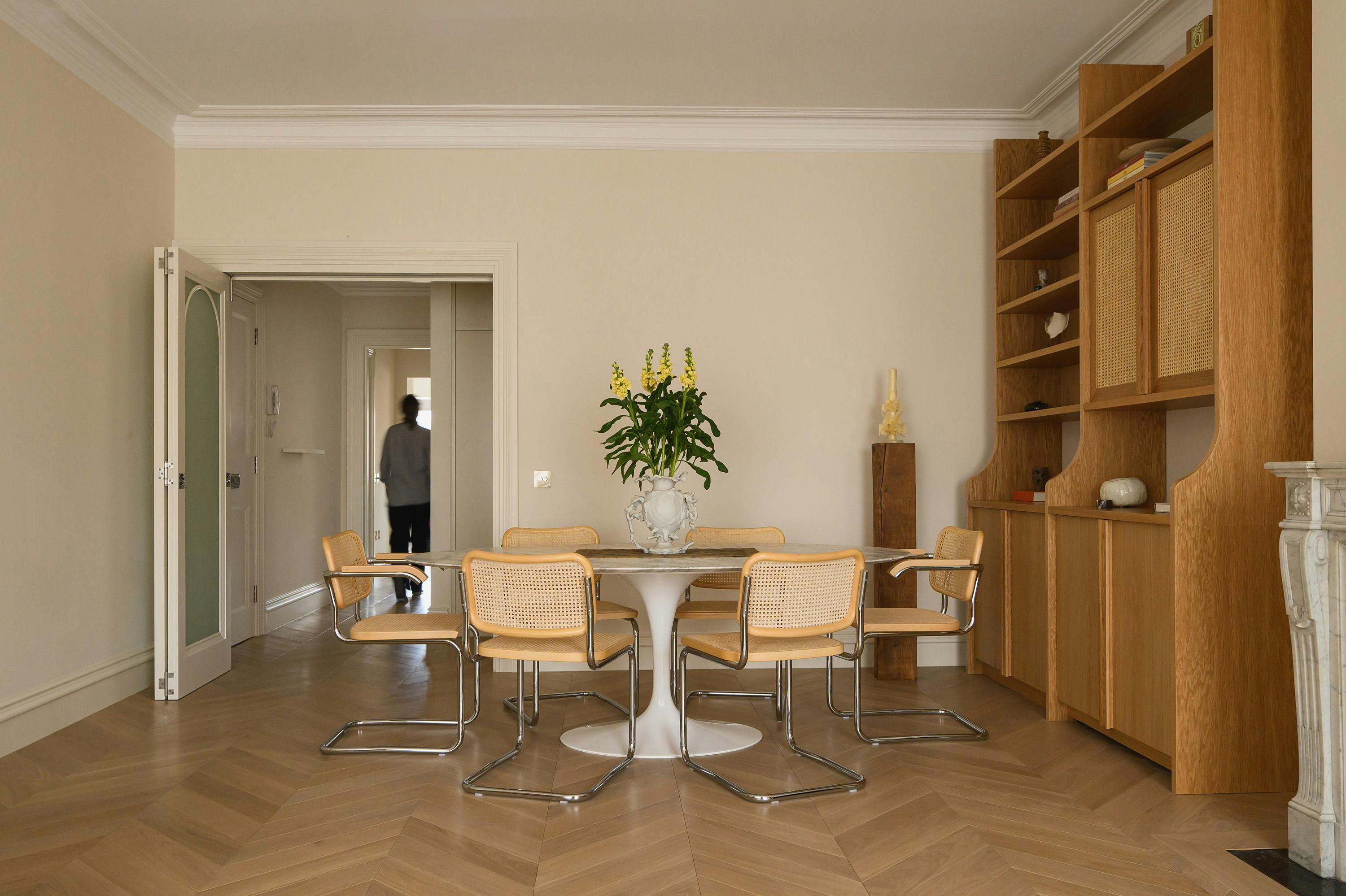 A modern dining area with a round marble-topped table and six rattan-backed chairs with chrome frames, arranged on a herringbone-patterned wooden floor. A decorative white vase with green foliage and yellow flowers serves as a centrepiece. To the right, a custom-built wooden shelving unit with open shelves and woven rattan cabinet doors displays books, ceramics, and sculptural decor. A white marble fireplace is partially visible on the far right. In the background, an open doorway leads to a hallway where a person in casual attire is walking away, adding a dynamic element to the serene, neutral-toned space.