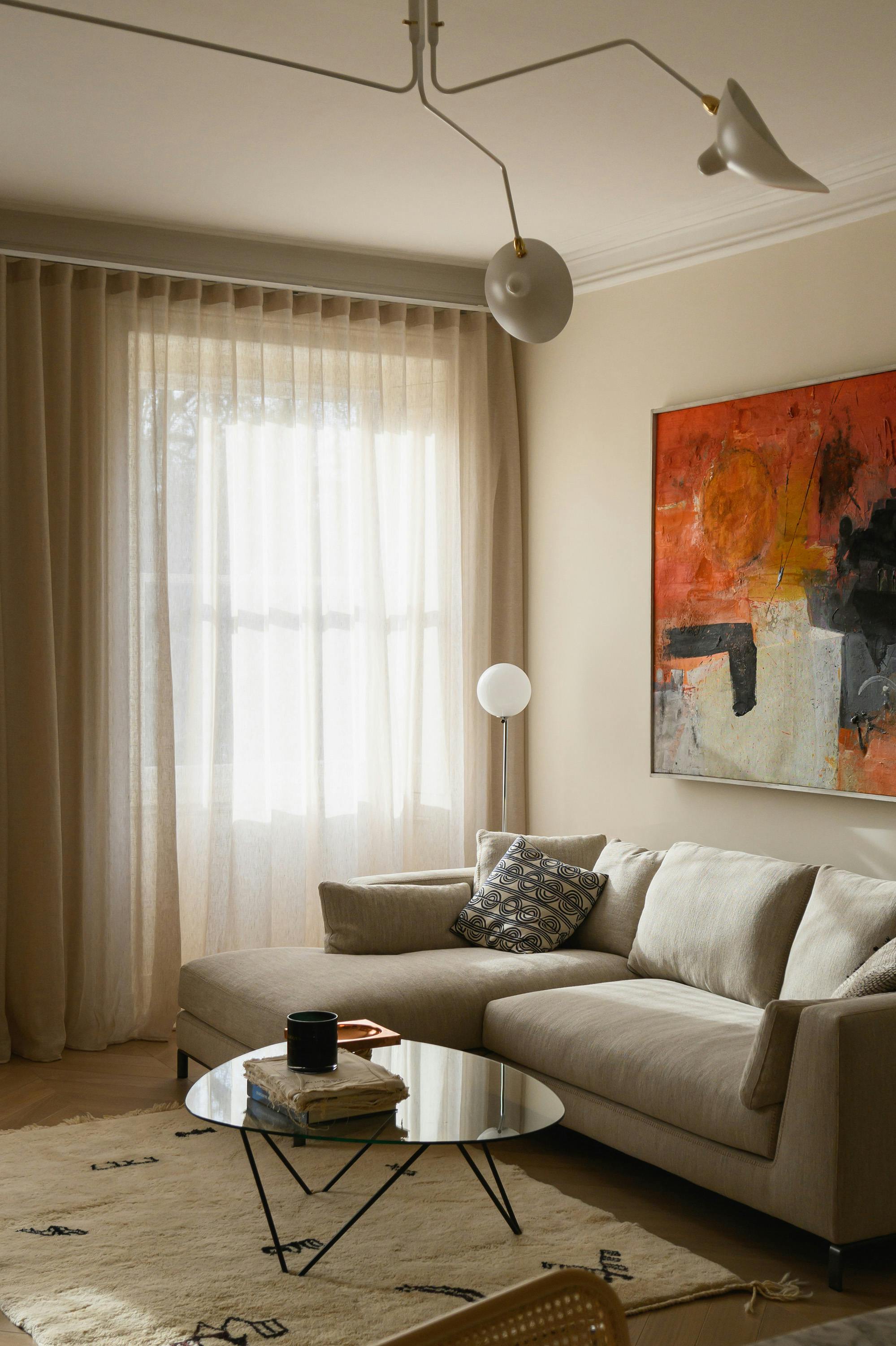 A cozy and elegantly designed living room featuring a neutral-toned L-shaped sofa adorned with patterned and textured cushions. A modern glass coffee table with a black metal frame sits atop a cream-colored rug with abstract black markings, holding a stack of books, a dark ceramic mug, and a decorative tray. Behind the sofa, an abstract painting with bold orange, black, and white tones adds vibrancy to the space. Soft, floor-length beige curtains cover a large window, allowing diffused natural light to enter. A contemporary white ceiling light fixture with sculptural shades hangs above, complementing the modern aesthetic of the space.