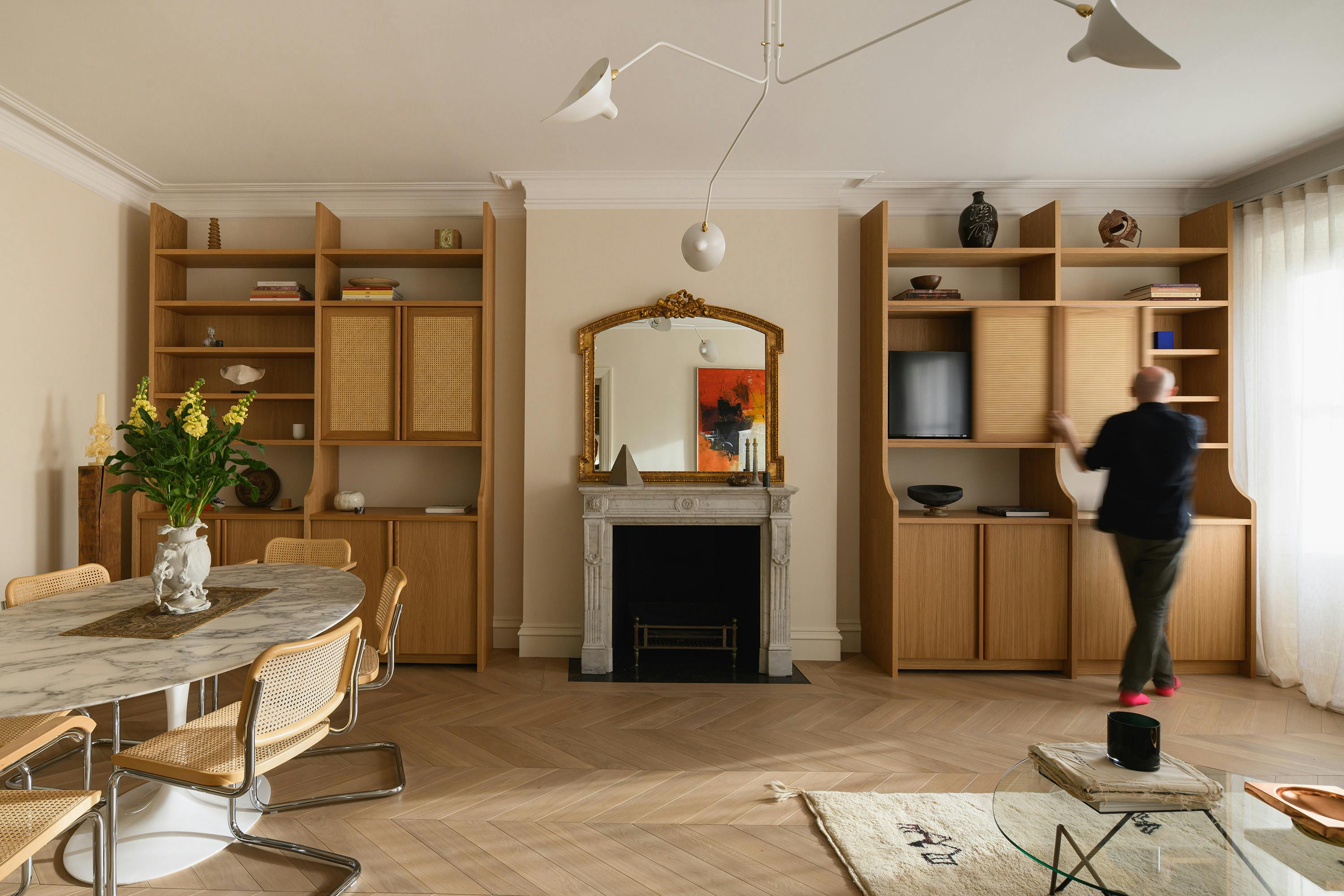 A stylish dining and living space featuring a marble-top dining table surrounded by six woven cane and metal chairs. A large wooden bookshelf with open shelving and rattan-fronted cabinets spans the wall, filled with books, decorative objects, and a concealed television. A person dressed in dark clothing and pink socks is seen opening one of the cabinet doors. A grand fireplace with an ornate gilded mirror above it serves as a focal point, reflecting an abstract painting with bold orange and black tones. The room is illuminated by a sculptural white ceiling light, while a glass coffee table and a cream rug with black markings add warmth and texture to the space. Natural light filters through sheer curtains on the right.