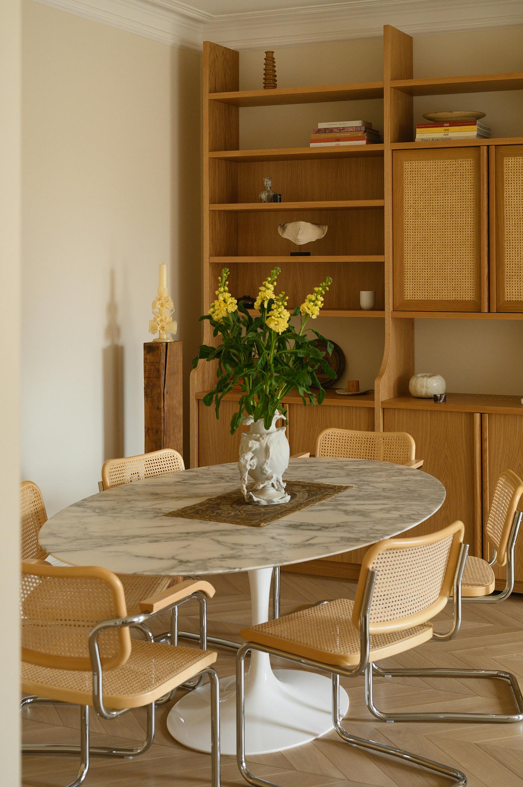 A close-up view of a dining area featuring a round marble table with a sculptural white vase holding fresh yellow flowers. Surrounding the table are woven cane and chrome dining chairs with a modern yet vintage aesthetic. Behind the dining setup, a custom-built wooden bookshelf with rattan-fronted cabinets is neatly styled with books, ceramics, and sculptural objects. A wooden pedestal holding a decorative candle stands against the neutral-toned wall. The room’s warm wood tones, natural textures, and soft lighting create an inviting and sophisticated atmosphere.