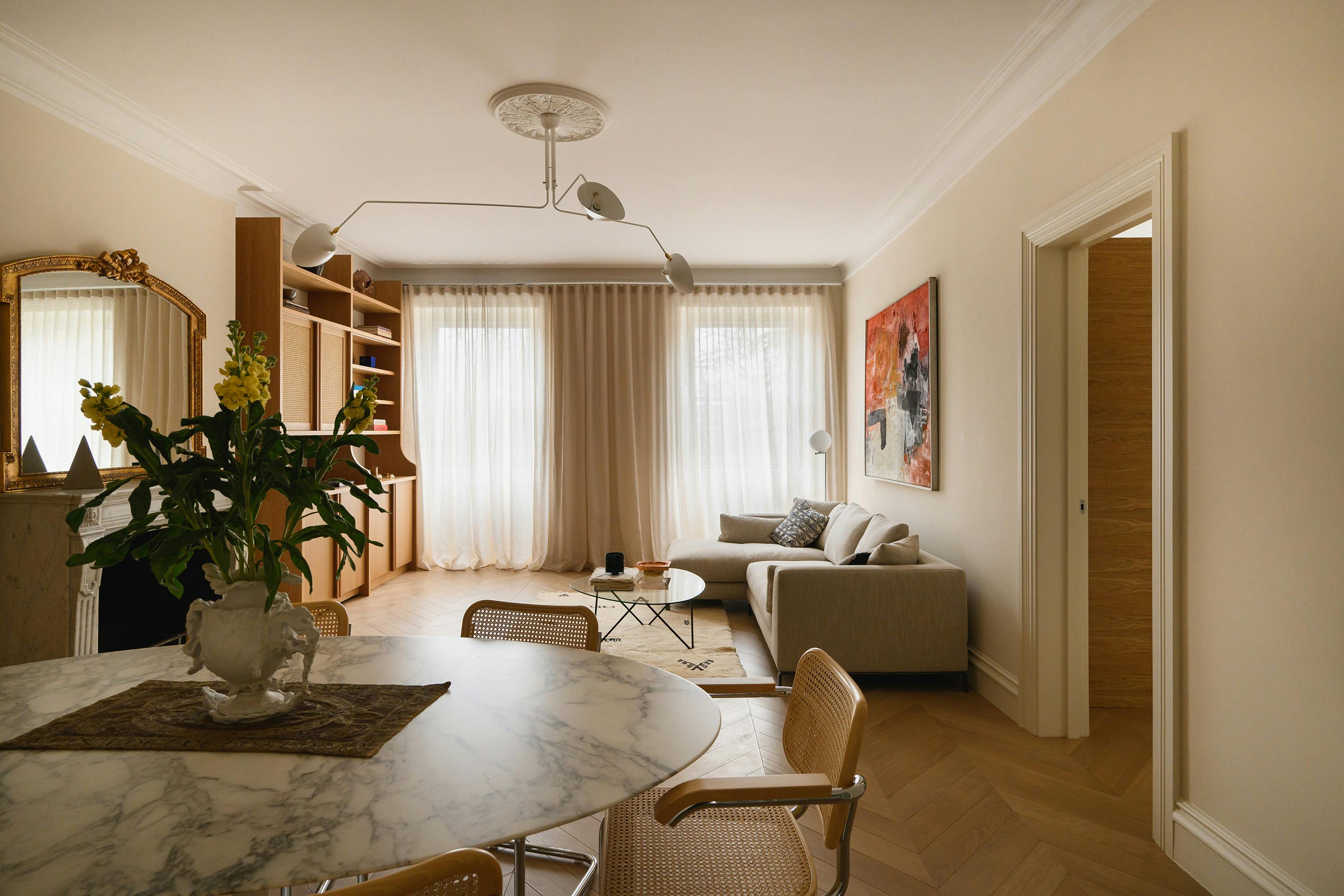 A stylish open-plan living and dining space with a neutral colour palette. In the foreground, a marble dining table with woven cane chairs is adorned with a sculptural white vase holding fresh yellow flowers. The dining area transitions into a cozy lounge featuring a beige L-shaped sofa, a glass coffee table, and a textured rug. A bold abstract painting with red and black hues hangs on the wall, adding contrast. Soft, sheer curtains frame large windows, allowing natural light to filter in. A modern, sculptural ceiling light fixture and a decorative gold-framed mirror above a classic fireplace add elegance to the space.