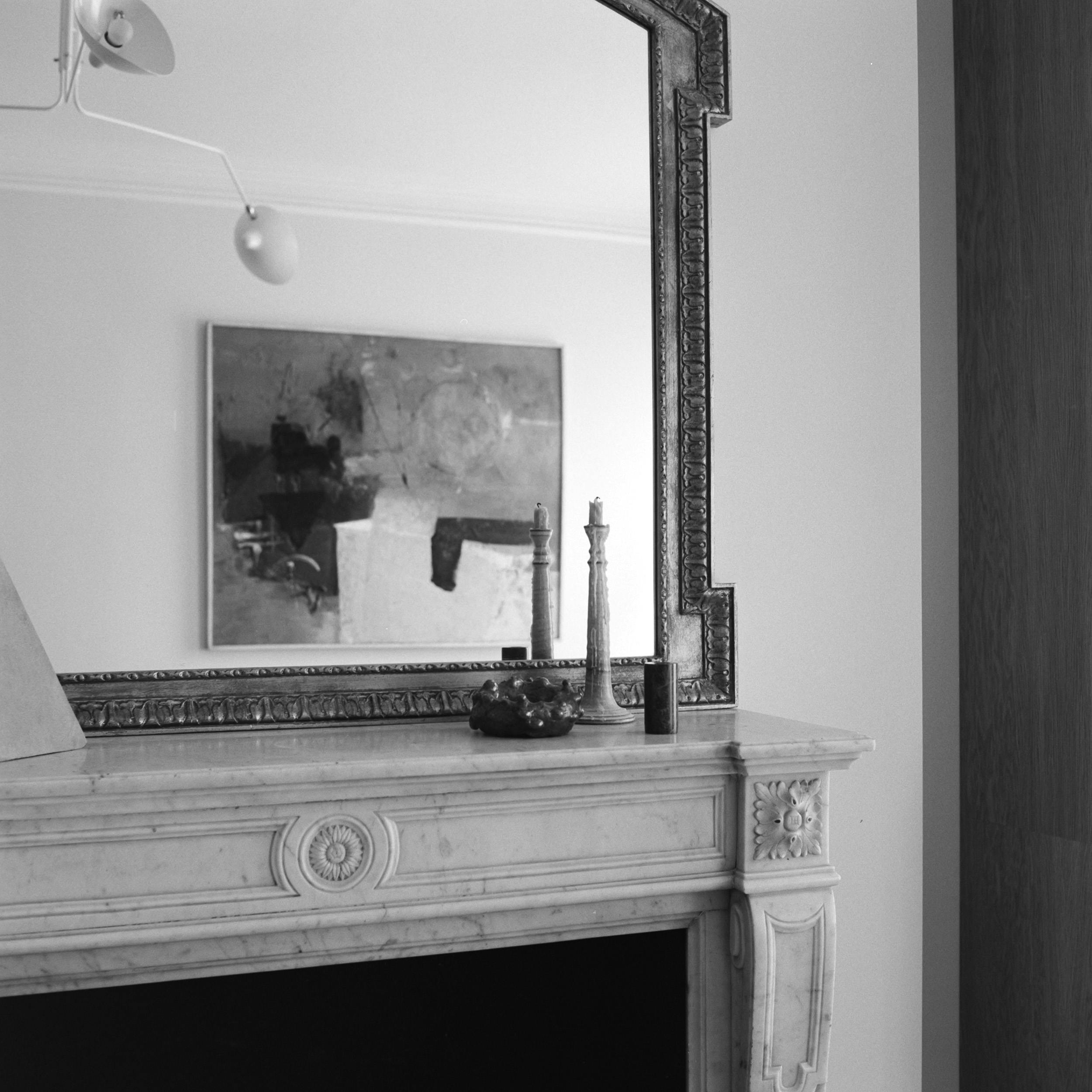 Black and white, angled close-up of a decorative fireplace and mirror. The light-coloured stone mantel, possibly marble, has intricate carvings including a rosette and floral designs. The firebox is dark and rectangular. Above, a large, ornate framed mirror reflects part of the room, including a framed abstract painting in grey and black. The mirror's thick frame has a geometric inner edge. On the mantel are two slender candlesticks and a small, dark textured object. A modern wall lamp with a white shade is in the upper left, lighting the wall and mirror. The right edge shows part of a dark wood panel or door. The image emphasizes the architectural and decorative details.