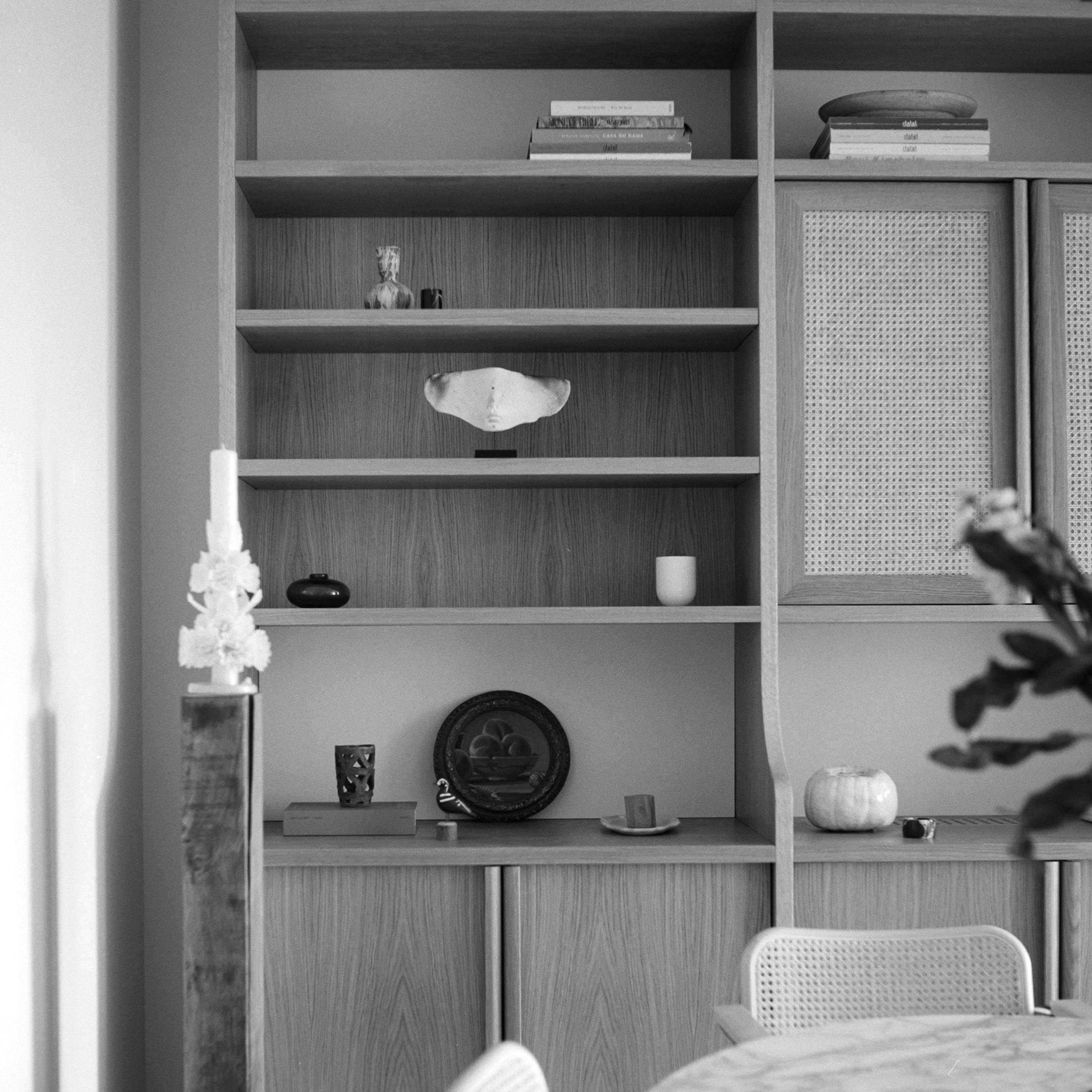 Black and white, angled close-up of a light-wood built-in bookshelf. The shelves, of varying heights, display decorative objects, books, and small sculptures. Cane-panelled cabinet doors are on the right. A round wooden table and white wicker chair are partially visible in the foreground. A plant and a tall, white decorative object are also present. The image shows a modern, minimalist interior design.