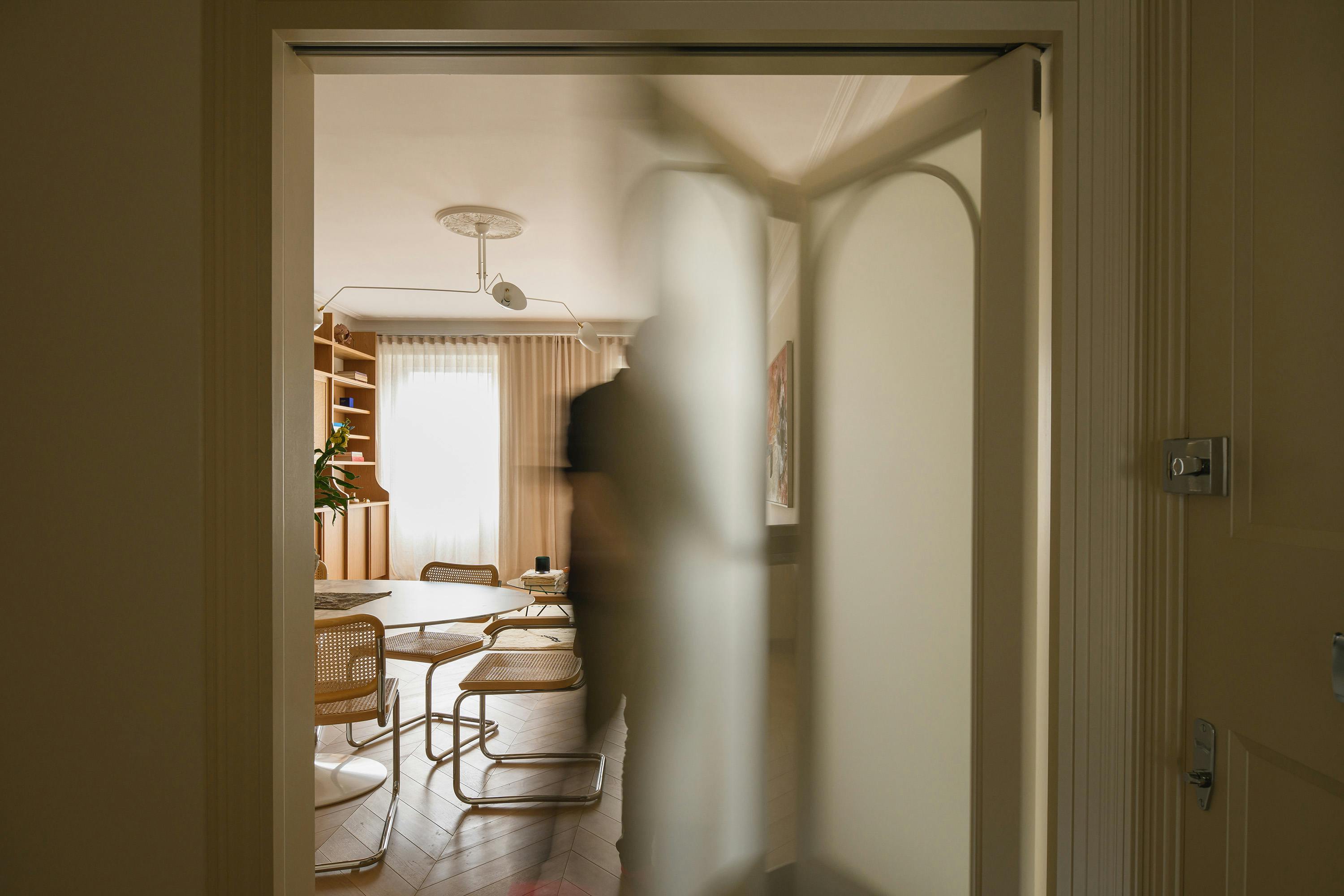 A slightly angled, eye-level photograph of a doorway leading into a brightly lit room. The doorway is framed by a white doorframe with a panelled design. A frosted or textured glass door is partially open, blurring the view into the room. Inside, a dining area is visible with a round table, wooden chairs with woven seats, and a built-in bookshelf against the far wall. A person's blurred silhouette is visible through the glass door, suggesting movement. A modern, multi-armed light fixture hangs from the ceiling. The room is well-lit, and the overall colour palette is warm and neutral.