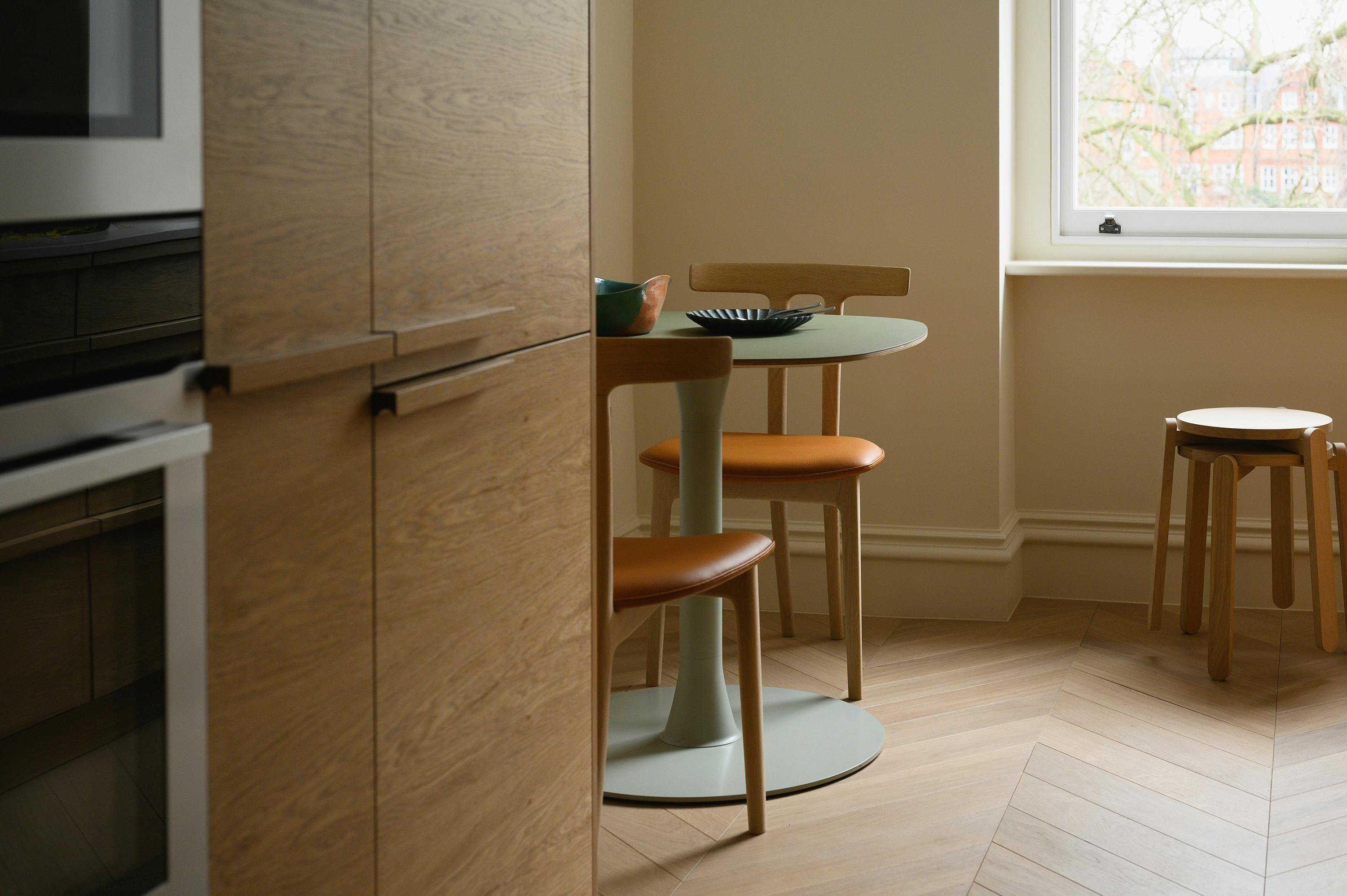 A slightly angled, eye-level photograph of a kitchen corner. On the left, light wood cabinets with horizontal grain are visible, featuring long, thin metal handles. A built-in oven with a white trim is partially visible. In the centre, a small round table with a light green top and a light green pedestal base stands on a light wood herringbone floor. Two wooden chairs with tan leather seats are tucked under the table. On the right, a window with a view of trees and buildings is visible, and a small wooden stool with a round seat stands near the wall. The walls are painted a warm beige colour. The lighting in the room is natural and bright