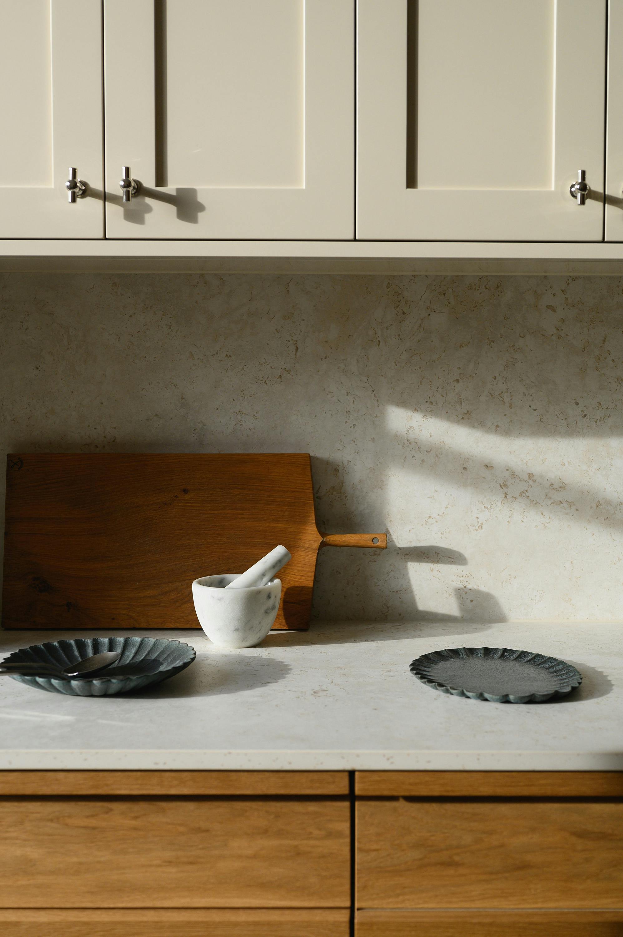 A close-up view of a dining area featuring a round marble table with a veined pattern and woven cane chairs with a light wood frame and chrome detailing. The chairs have a mid-century modern aesthetic, with a curved backrest and armrests. In the background, an open doorway leads to a kitchen with wooden cabinetry, a stainless steel faucet, and a light-coloured backsplash. The room is illuminated by natural light, highlighting the warm tones of the wooden flooring and furniture.