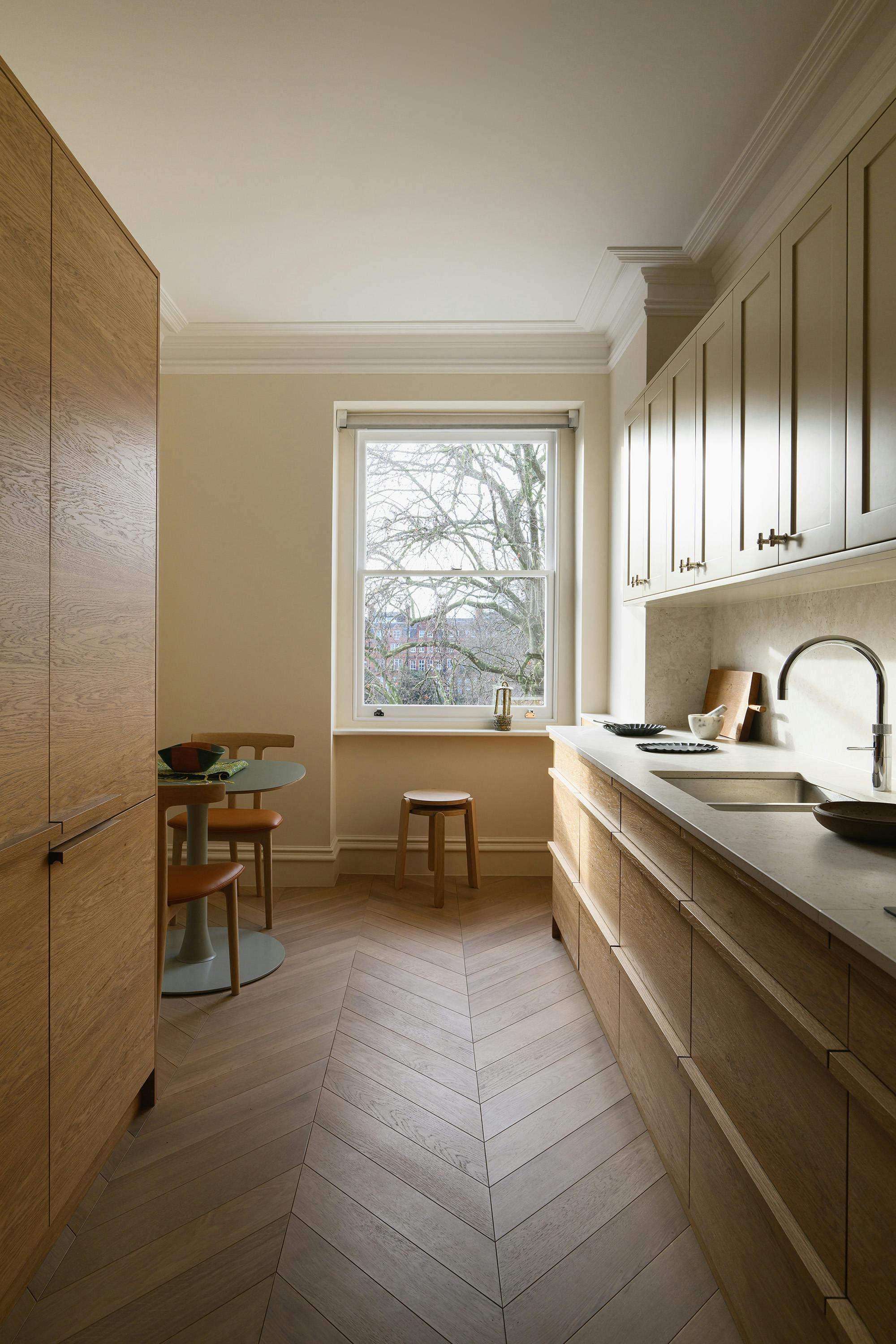 A slightly elevated, straight-on photograph of a narrow kitchen. The kitchen features light-coloured upper cabinets with traditional panelling and silver knobs, and medium-toned wood lower cabinets with a horizontal grain. A light-coloured countertop with a speckled pattern runs along the right side of the kitchen, featuring a stainless steel sink with a curved faucet. A window with a white frame and a view of trees is centred at the far end of the kitchen. A small round table with two chairs is visible on the left side, along with a tall, matching wooden cabinet. A small wooden stool is placed near the window. The floor is a light-coloured herringbone pattern. The walls are painted a warm beige colour. The overall aesthetic is modern, clean, and bright, with a focus on natural light.