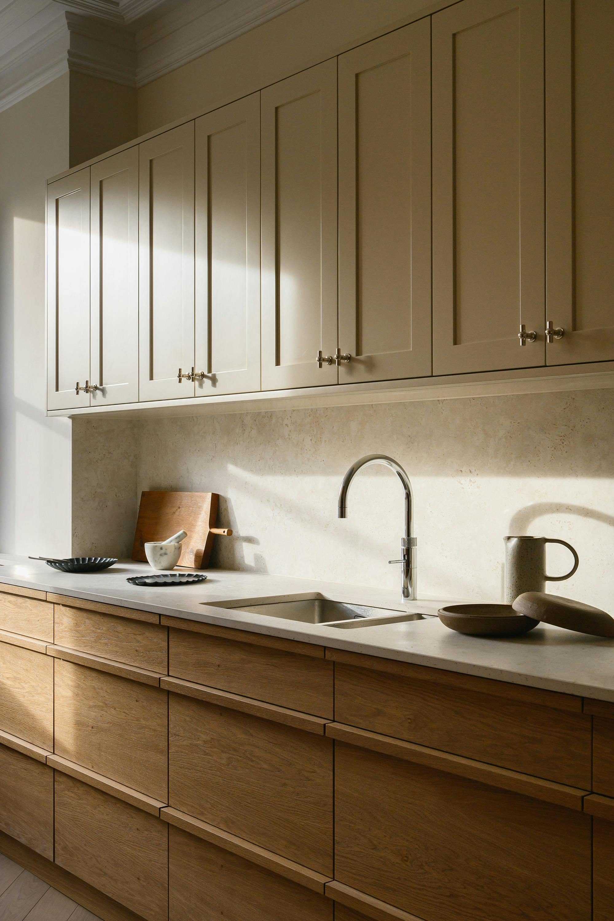 slightly angled, eye-level photograph of a kitchen featuring a combination of light and medium-toned wood cabinets. The upper cabinets are painted a light, creamy colour with traditional panelling and small, silver knobs. Below the upper cabinets is a light-coloured countertop with a speckled or textured pattern, resembling stone. A stainless steel sink with a curved faucet is cantered on the countertop. On the left side of the countertop, a dark grey textured plate, a light-coloured stone mortar and pestle, and a wooden cutting board are visible. On the right side, a light-coloured pitcher and a wooden spoon are placed. The lower cabinets are made of a medium-toned wood with a horizontal grain. Sunlight casts shadows across the upper cabinets and countertop, highlighting the textures and colours. The overall aesthetic is modern, clean, and natural.