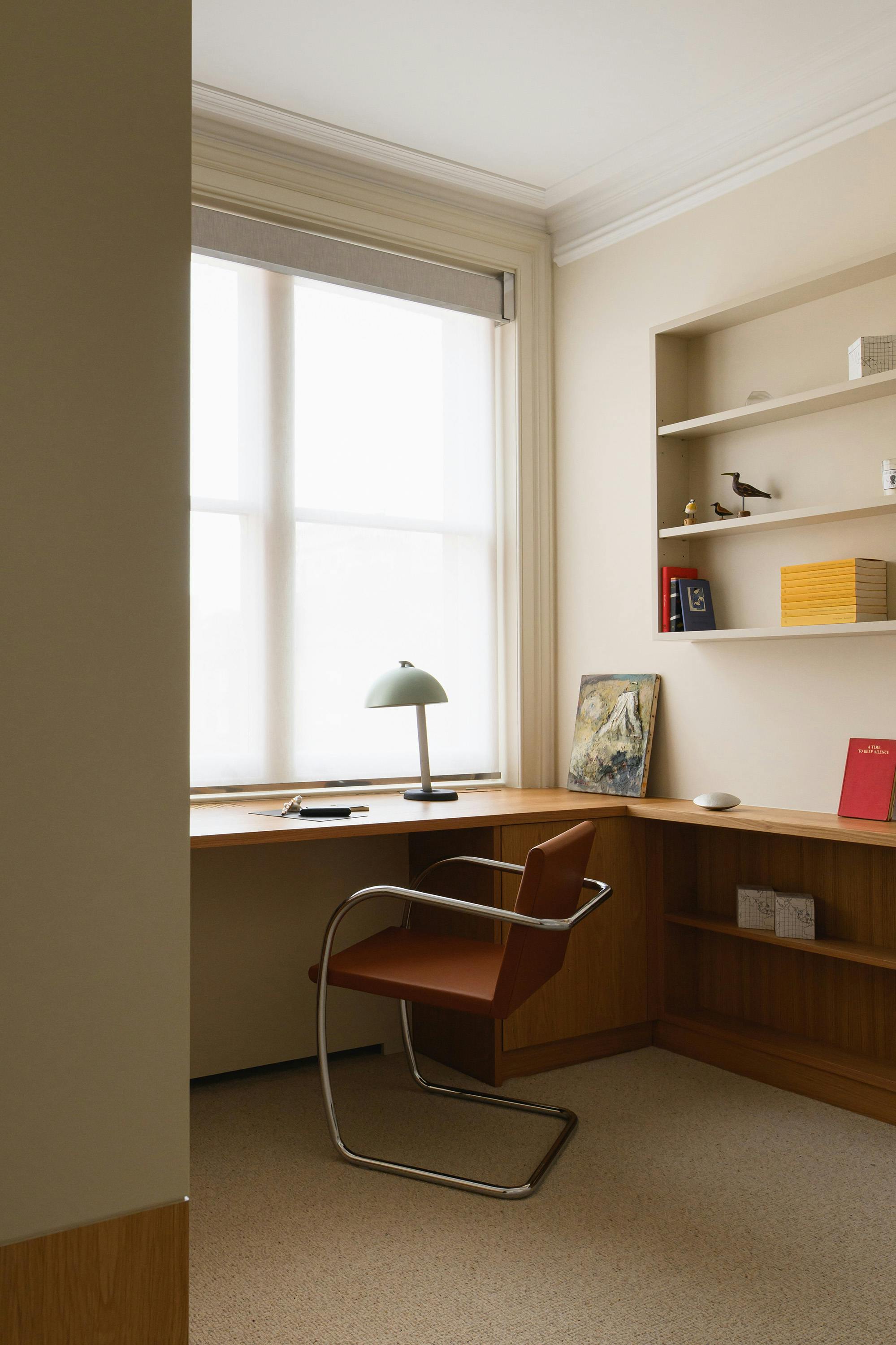A slightly elevated, straight-on photograph of a corner office space. On the left, a large window with a white blind is visible, with a wooden desk running along the wall underneath. A light green lamp and a small black object are placed on the desk. In the centre, a tan leather chair with a chrome cantilever frame (Brno chair) is positioned in front of the desk. On the right, built-in wooden shelves with decorative items are visible, including books, a painting, and small figurines. The walls are painted a light, neutral colour. The floor is covered with a light-coloured carpet with subtle speckles. The overall aesthetic is modern and minimalist, with a focus on clean lines and natural light. A portion of a dark wood panel is visible in the lower left corner.