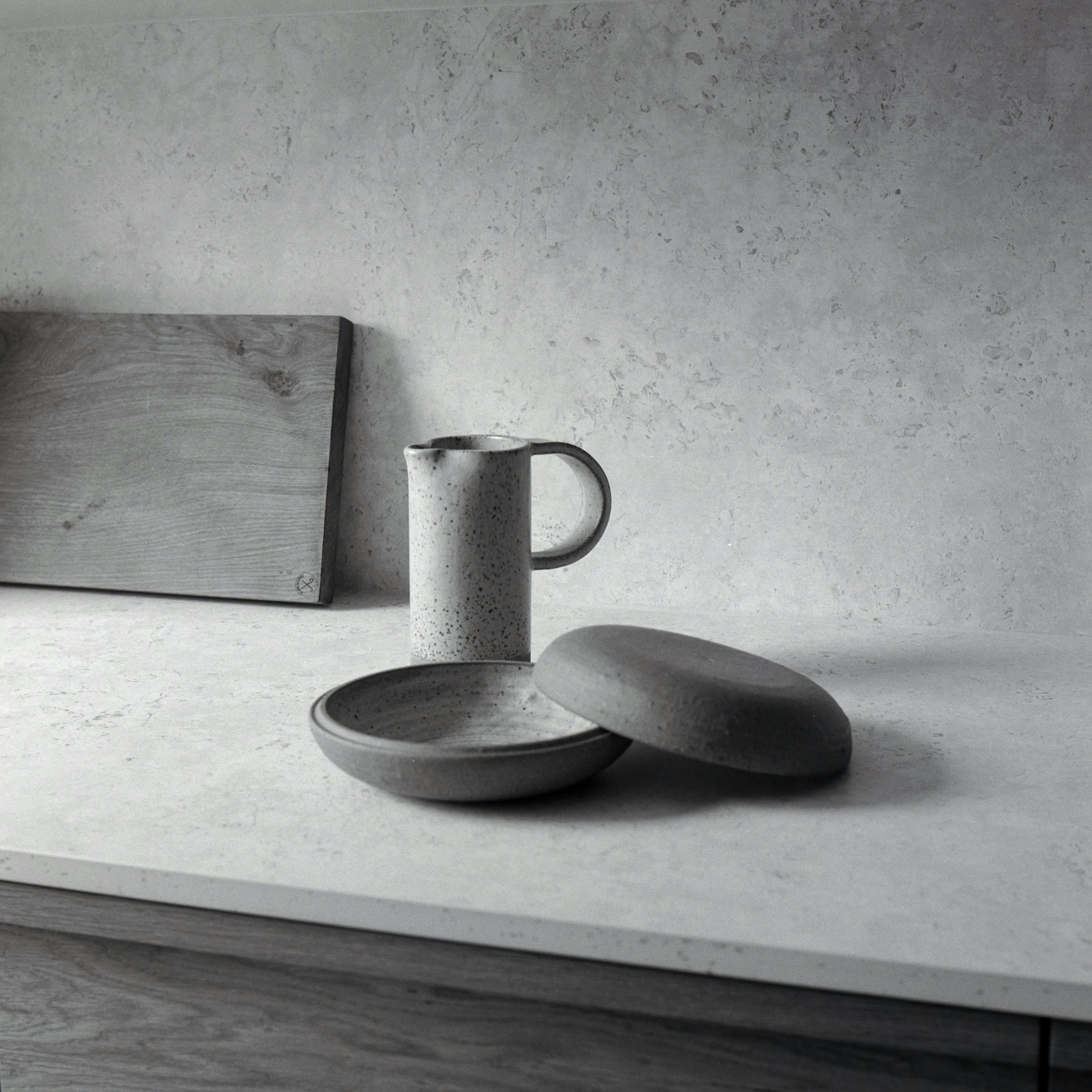 A close-up, monochromatic photograph of a kitchen countertop. The countertop is light-coloured with a subtle speckled or textured pattern, resembling stone. On the countertop, from left to right, are: a rectangular wooden cutting board, a light-coloured ceramic pitcher with a handle, and a dark grey ceramic bowl with a matching lid placed beside it. The image is in shades of grey, highlighting the textures and shapes of the objects. The lower edge of the image shows a portion of a dark wood cabinet.