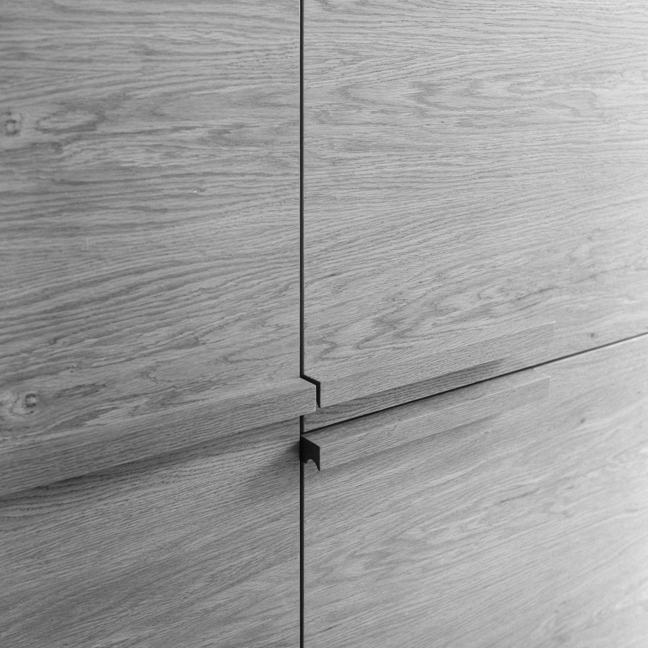 A close-up, monochromatic photograph of a light wood cabinet surface. The image focuses on the wood grain and the seam between two cabinet doors. The wood shows a distinct horizontal grain pattern. A thin, dark line marks the seam between the doors. A short, rectangular handle is visible on the right door, extending slightly outward. The image is in shades of grey, emphasizing the texture and details of the wood and hardware.