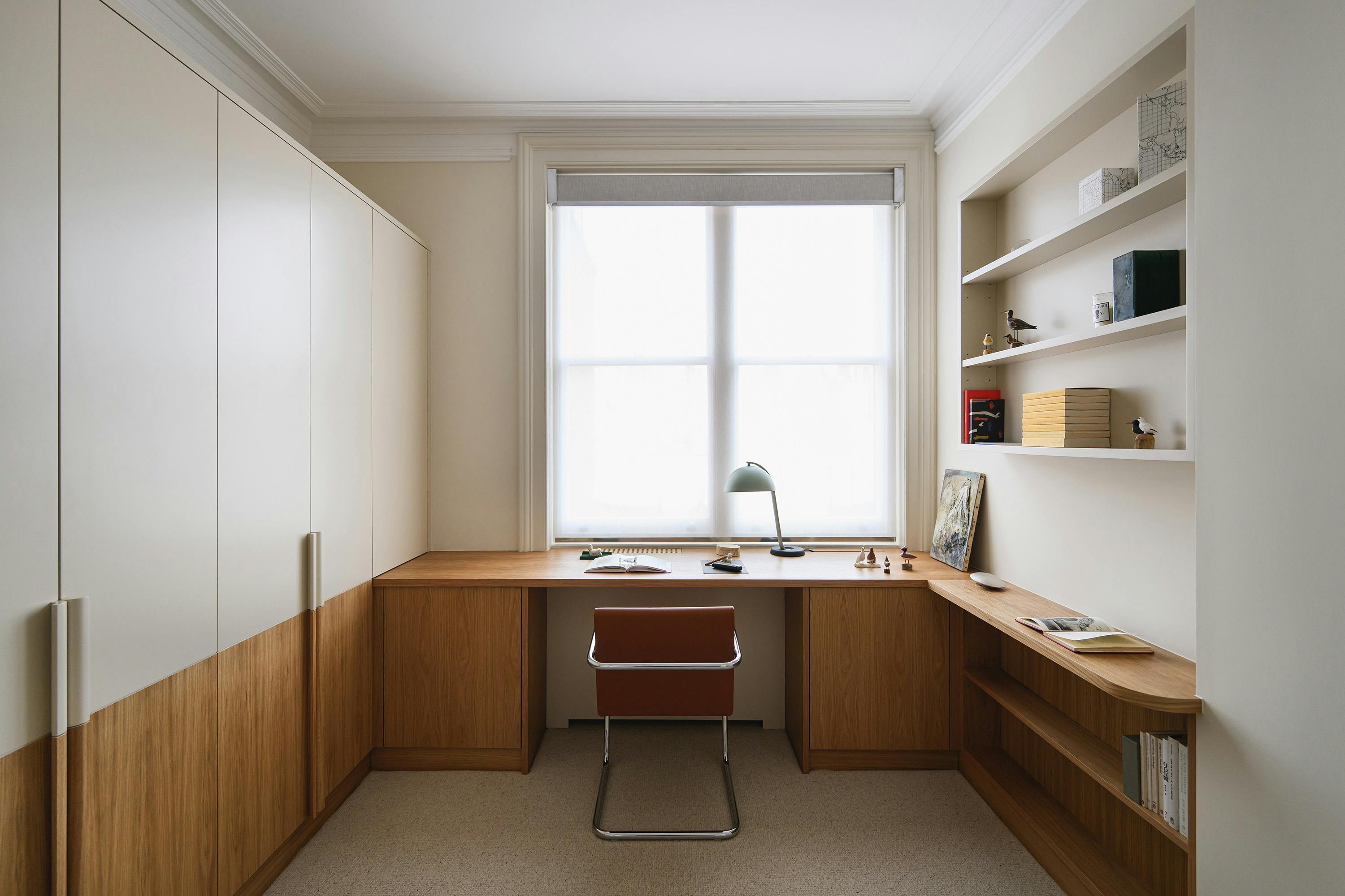 A slightly elevated, straight-on photograph of a small, well-organized home office. The office features a built-in wardrobe or cabinet unit on the left, combining light-coloured upper doors with medium-toned wood lower panels. A built-in wooden desk extends from the wardrobe to the right wall, creating a continuous workspace. A tan leather chair with a chrome cantilever frame (Brno chair) is positioned in front of the desk. A light green lamp and a book are placed on the desk. A window with a white blind is centred at the far end of the office, providing natural light. On the right wall, built-in shelves are visible, displaying books and decorative items. The floor is covered with a light-coloured carpet. The walls are painted a light, neutral colour. The overall aesthetic is modern, minimalist, and functional.