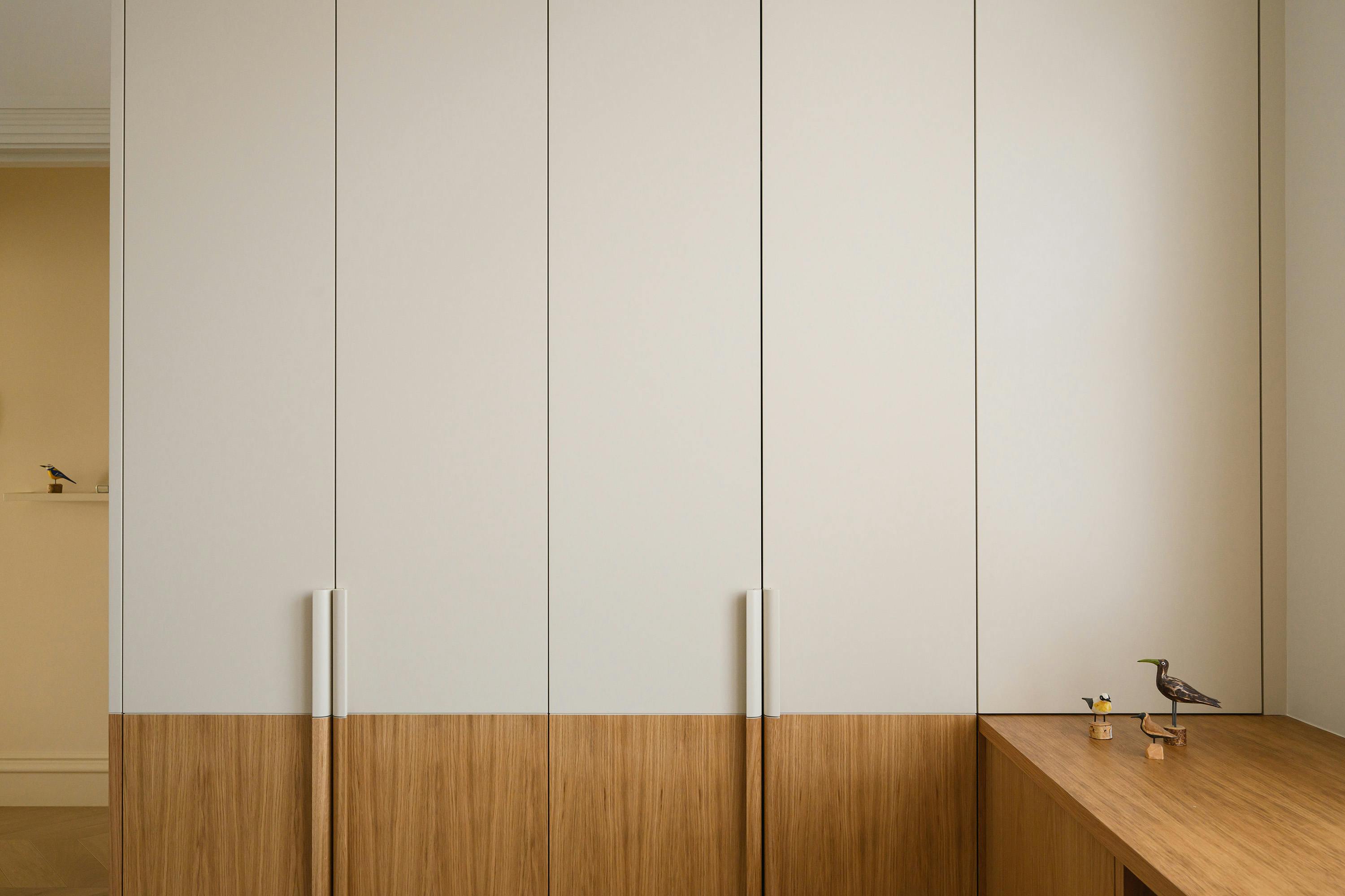 A straight-on, eye-level photograph of a built-in wardrobe or cabinet unit. The unit features a combination of light and medium-toned wood. The upper section consists of four vertically panelled doors, painted a light, creamy colour. Each door has a long, thin, silver handle. The lower section features horizontally grained, medium-toned wood panels. On the right side, a matching wooden surface extends outward, creating a small shelf or countertop. Two small bird figurines, one dark and one light, are placed on this surface. A small shelf with a single bird figurine is visible on the left wall. The overall design is modern and minimalist with clean lines and a focus on natural materials.
