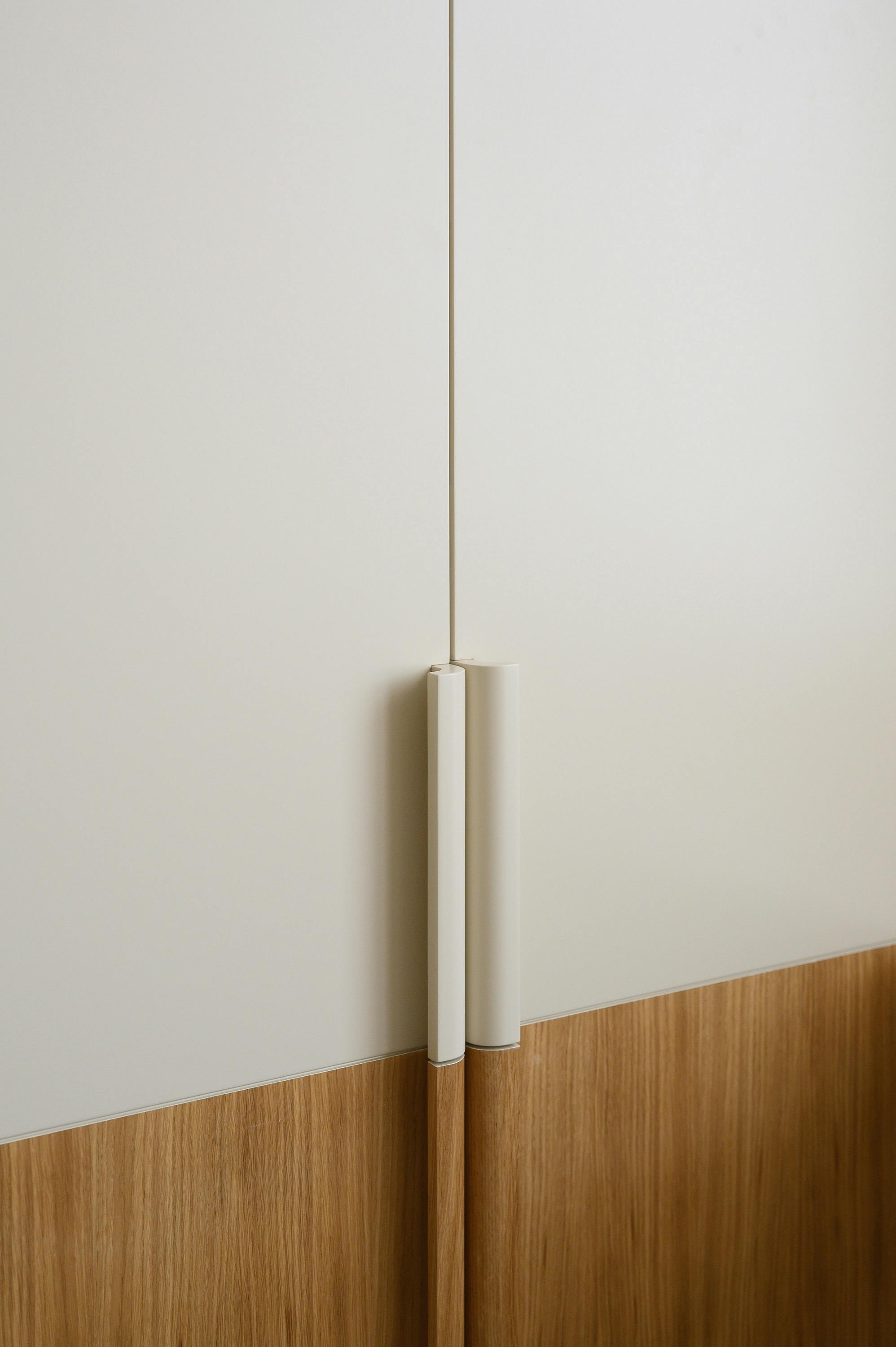 A slightly angled, eye-level photograph of a built-in wardrobe or cabinet unit in a corner. The unit features a combination of light and medium-toned wood. The upper section consists of vertically panelled doors, painted a light, creamy colour. A long, thin, silver handle is visible on one of the doors. The lower section features horizontally grained, medium-toned wood panels. On the right side, a matching wooden surface extends outward, creating a small shelf or countertop in the corner. Three small bird figurines, one dark and two light, are placed on this surface. The walls are painted a light, neutral colour. The overall design is modern and minimalist with clean lines and a focus on natural materials.