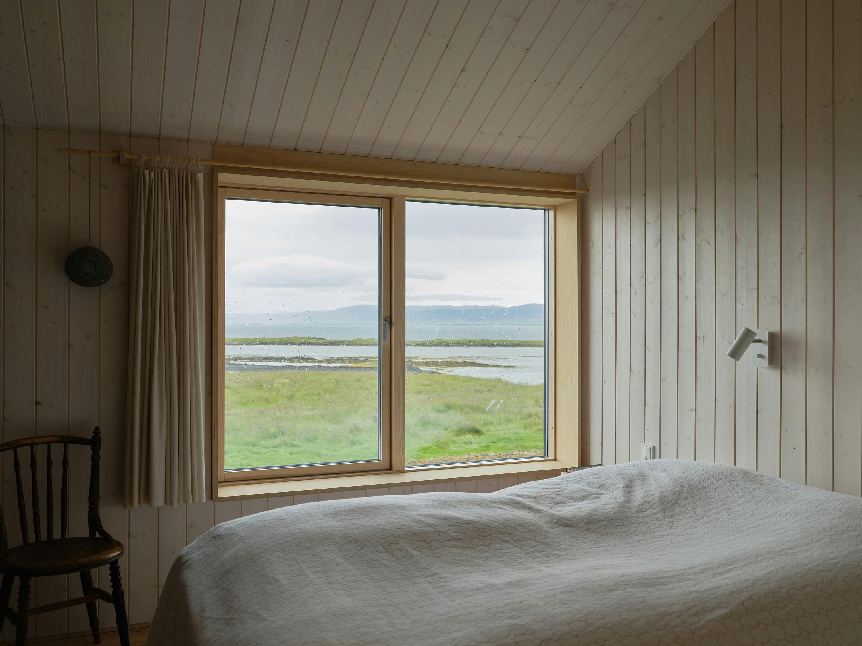 A slightly angled, eye-level photograph of a bedroom with light-coloured, vertically panelled wood walls and a sloped ceiling. A large rectangular window is centred on the far wall, offering a view of a grassy landscape, a body of water, and distant mountains. A light-coloured curtain is drawn to the left side of the window. A wooden chair with a dark finish is placed near the left wall. A bed with a light-coloured quilted cover is visible in the foreground, partially cropped. A small, white, angular light fixture is mounted on the upper right wall. The overall aesthetic is cozy and natural, with a focus on the view and simple materials