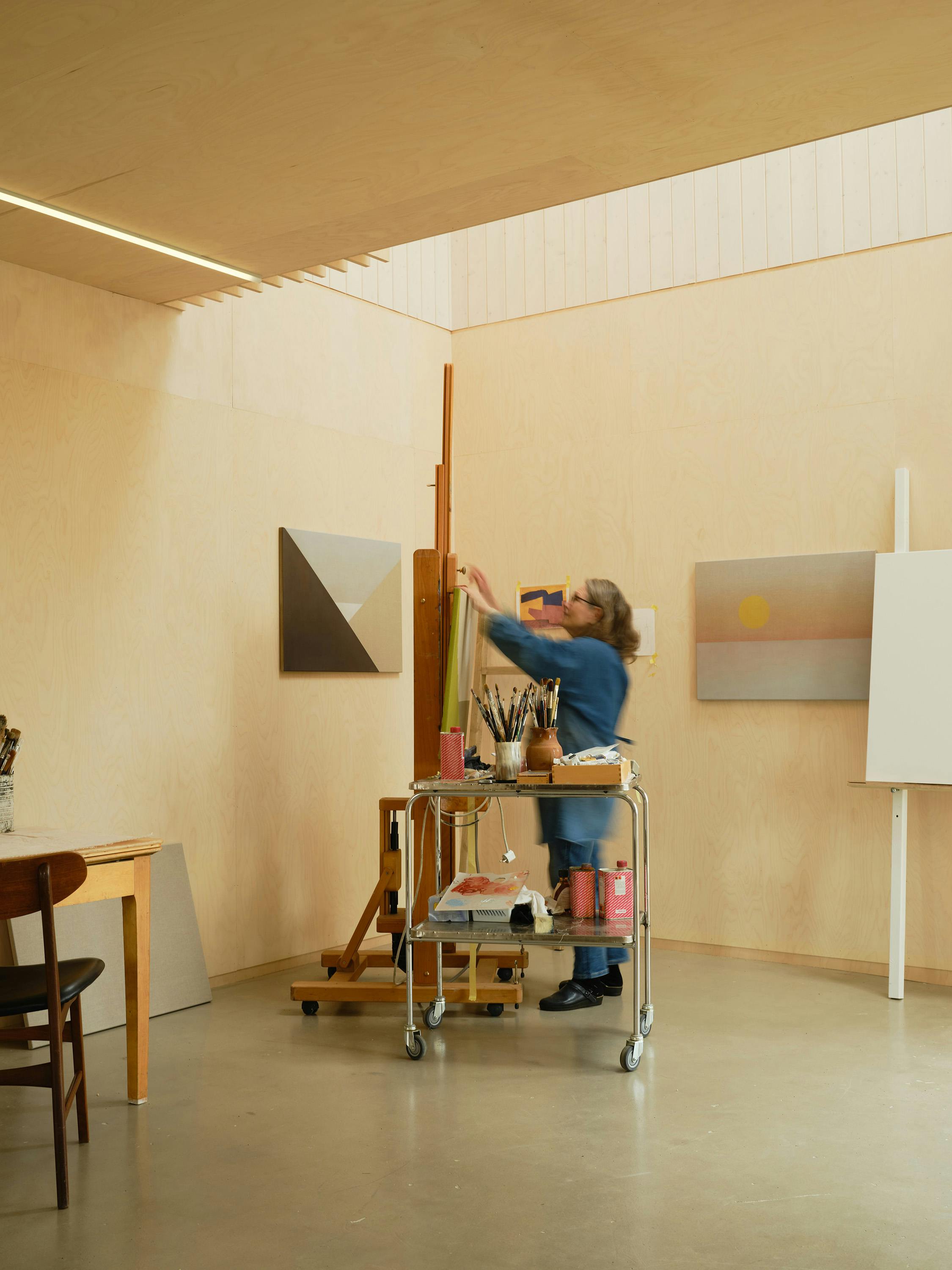 A female artist at work at an easel in a double height studio. The space is daylit with warm wooden walls and a polished concrete floor.
