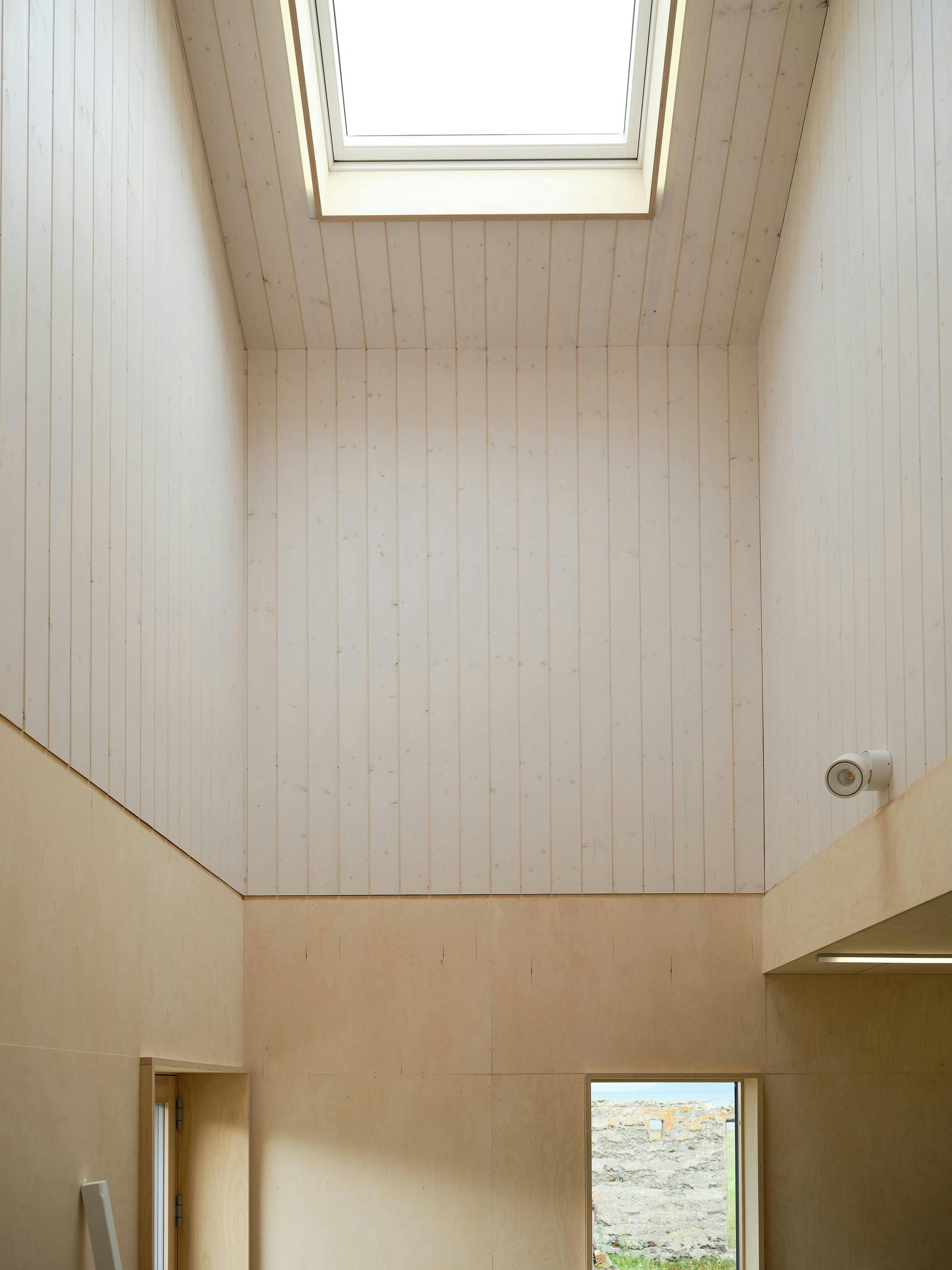 A straight-on, low-angle photograph of a tall, narrow room with a skylight. The room features light-coloured, vertically panelled wood walls and a light-coloured wood floor. A rectangular skylight is centred at the top of the image, allowing natural light to enter. A small, white, round light fixture is visible on the right wall. A doorway is visible on the left side of the room, and a window with a view of a rocky landscape is visible at the bottom of the image. The overall aesthetic is minimalist and bright, with a focus on natural materials and light.