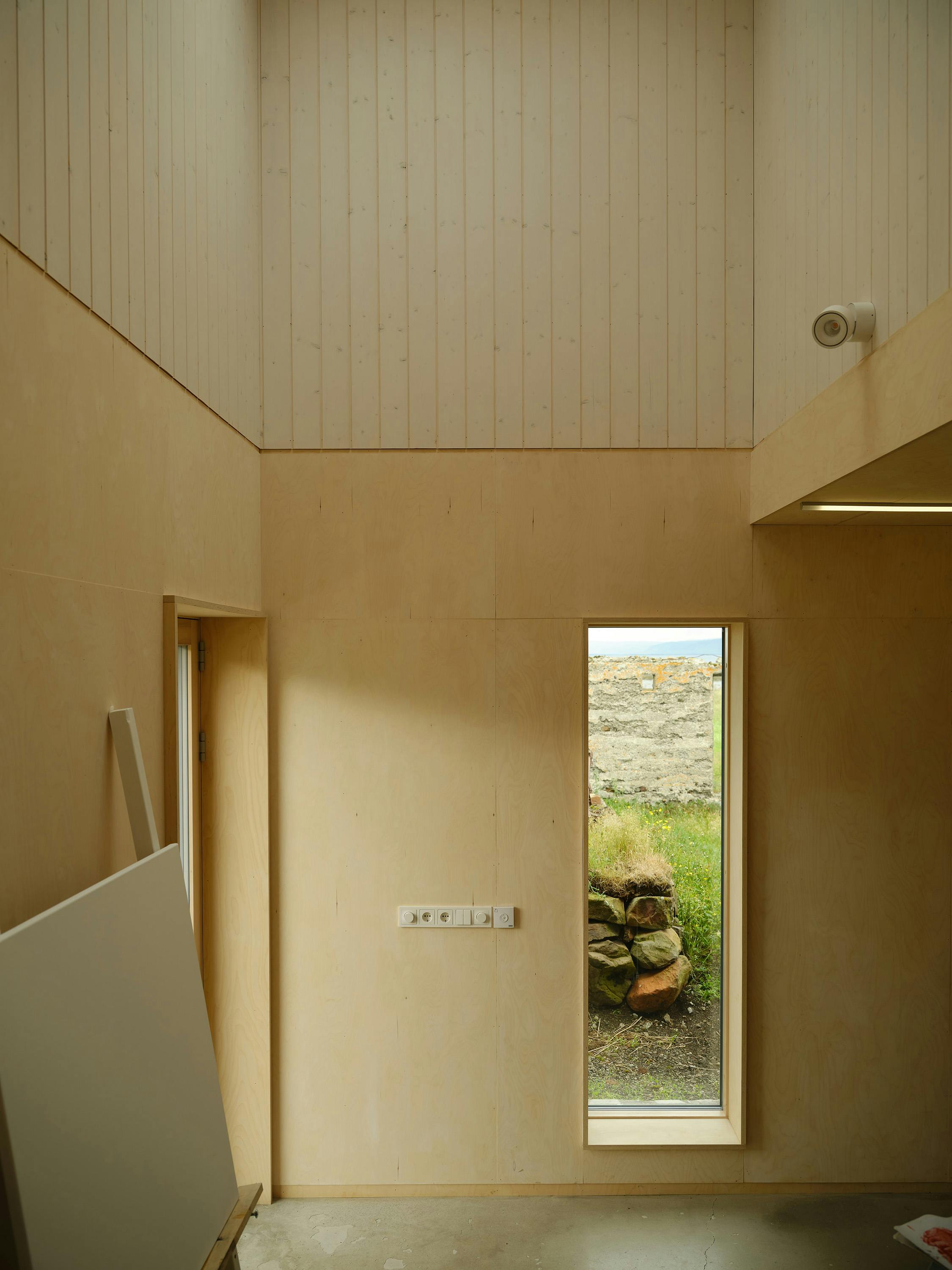 A slightly angled, eye-level photograph of a room with light-coloured wood walls and a concrete floor. The room features a combination of vertically panelled wood on the upper walls and smooth, light-coloured wood on the lower walls. A doorway is visible on the left side of the room, with a large white panel leaning against the wall. A rectangular window is centred on the far wall, offering a view of a rocky landscape with green grass and a pile of stones. A small, white, round light fixture is visible on the upper right wall. A long, thin light fixture is recessed into the ceiling on the right side. The overall aesthetic is minimalist and natural, with a focus on simple materials and the view outside.